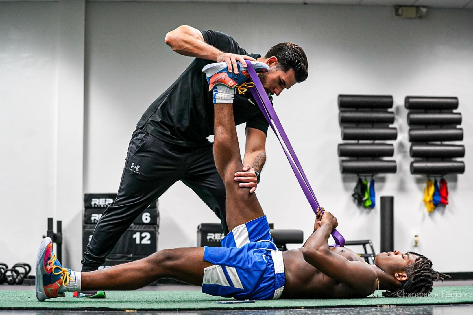 A man is stretching a man 's leg with a resistance band in a gym.