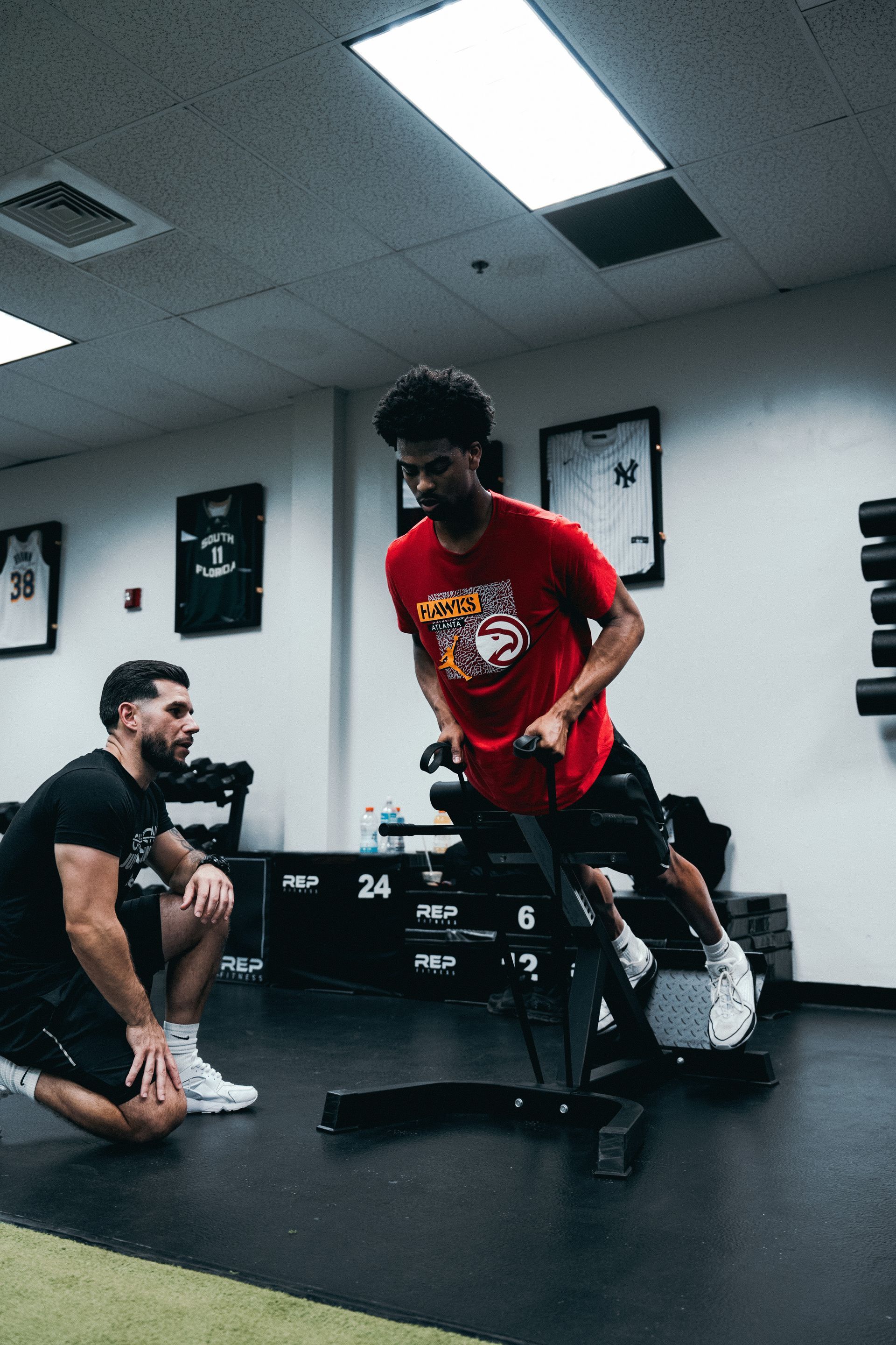 A man is kneeling down next to a man riding a bike in a gym.