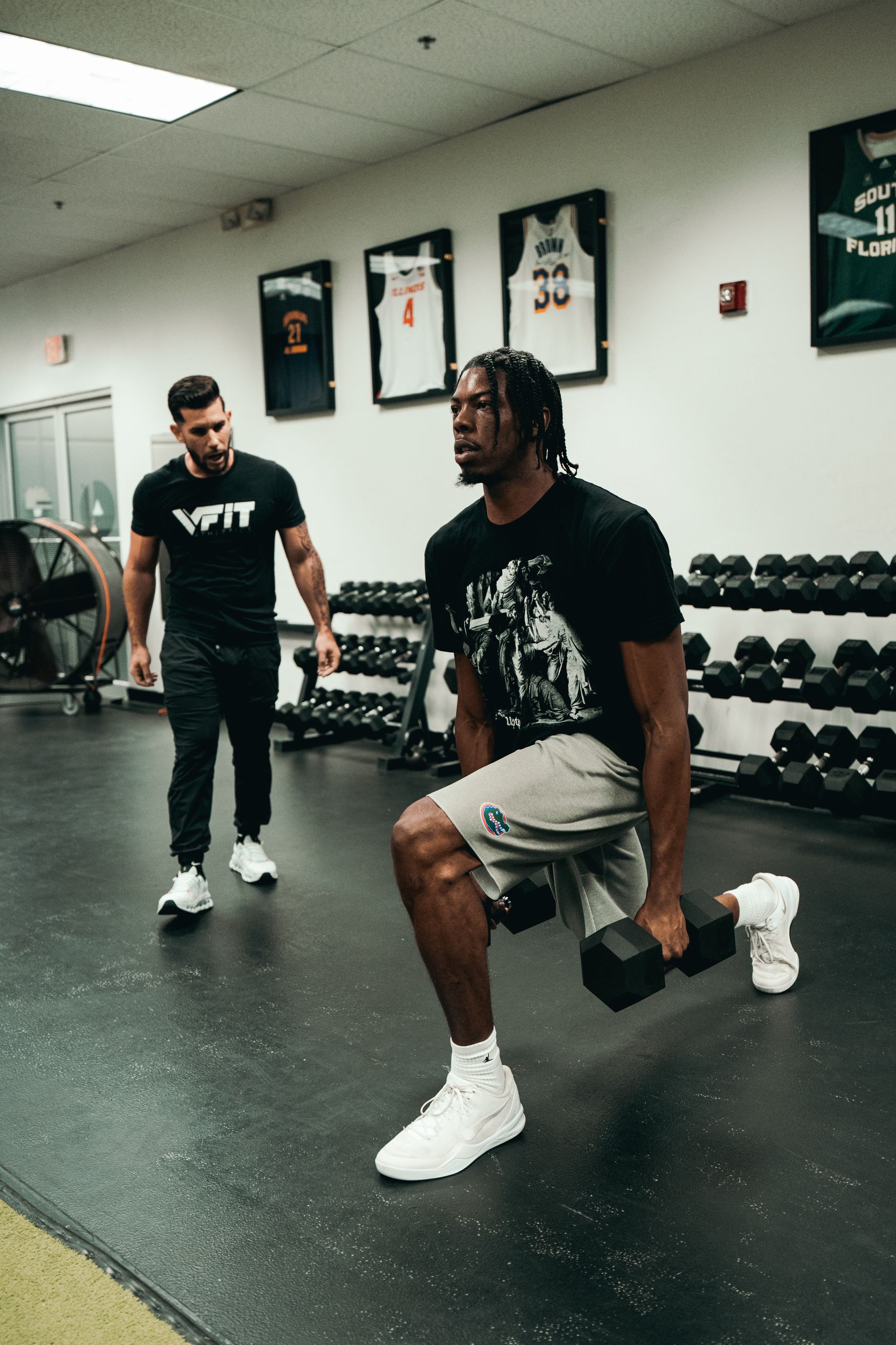 A man is squatting with a dumbbell in a gym while another man watches.