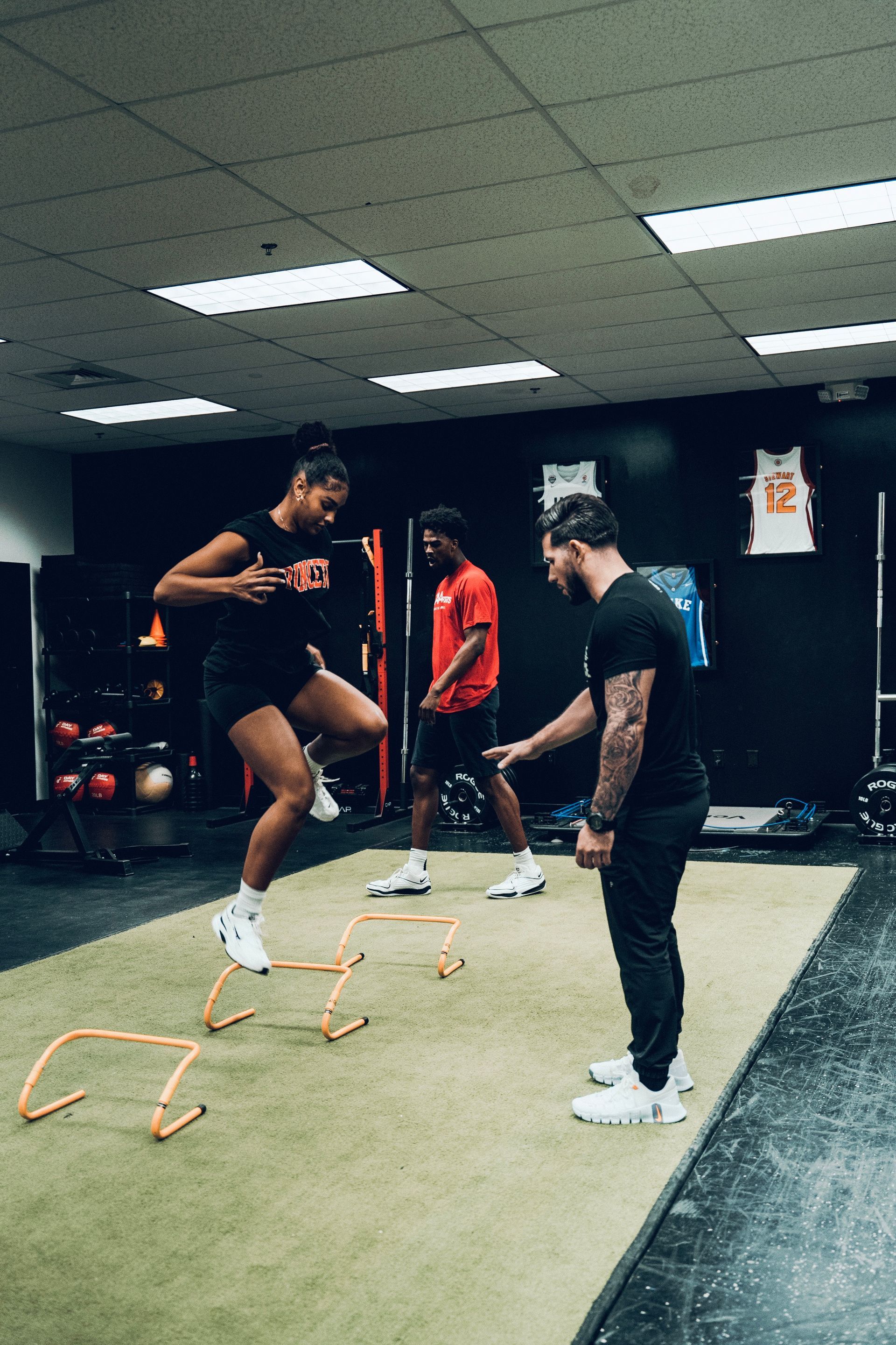 A man is standing next to a woman jumping over hurdles in a gym.