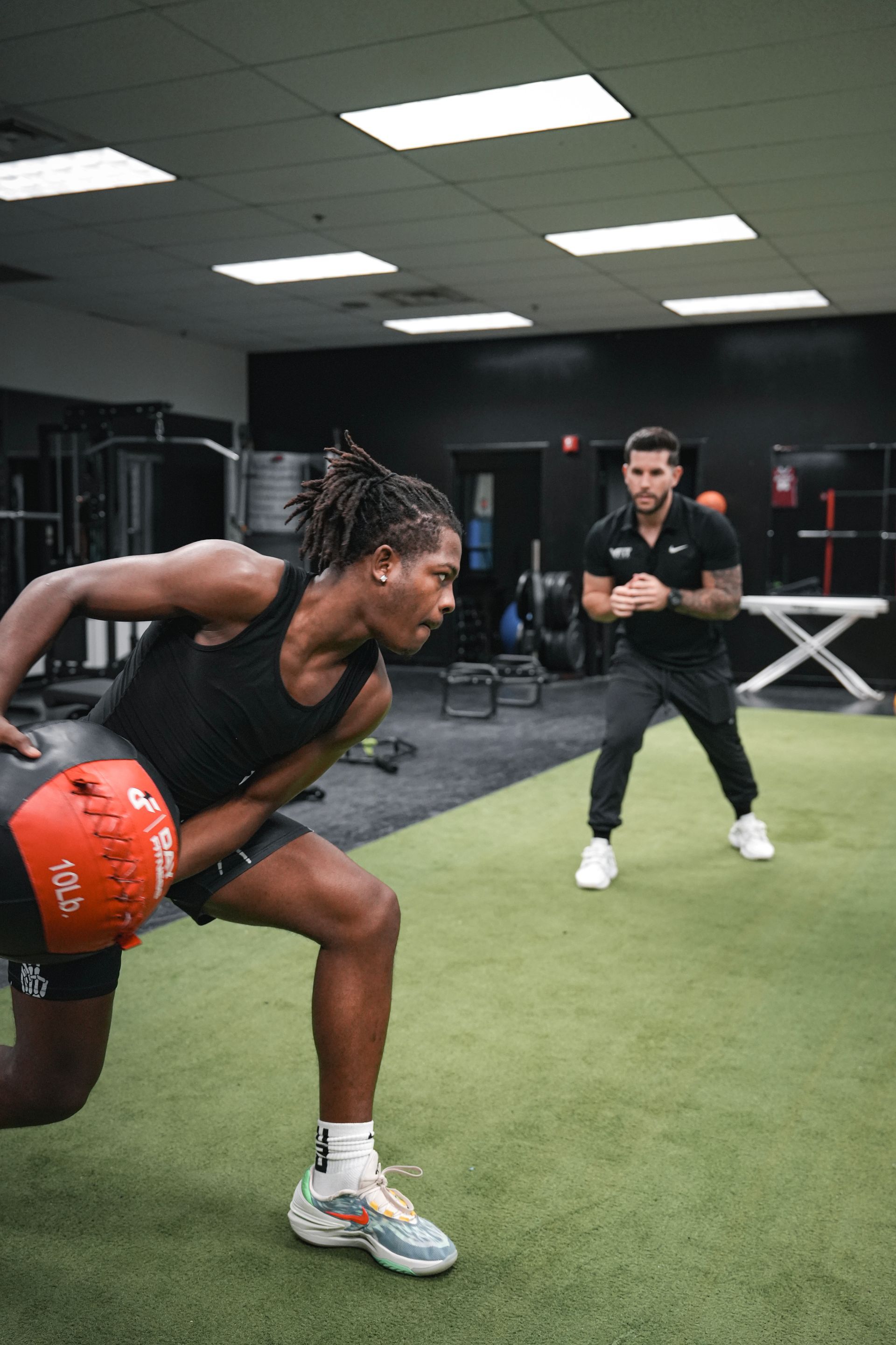 A man is squatting with a ball in a gym while another man watches.