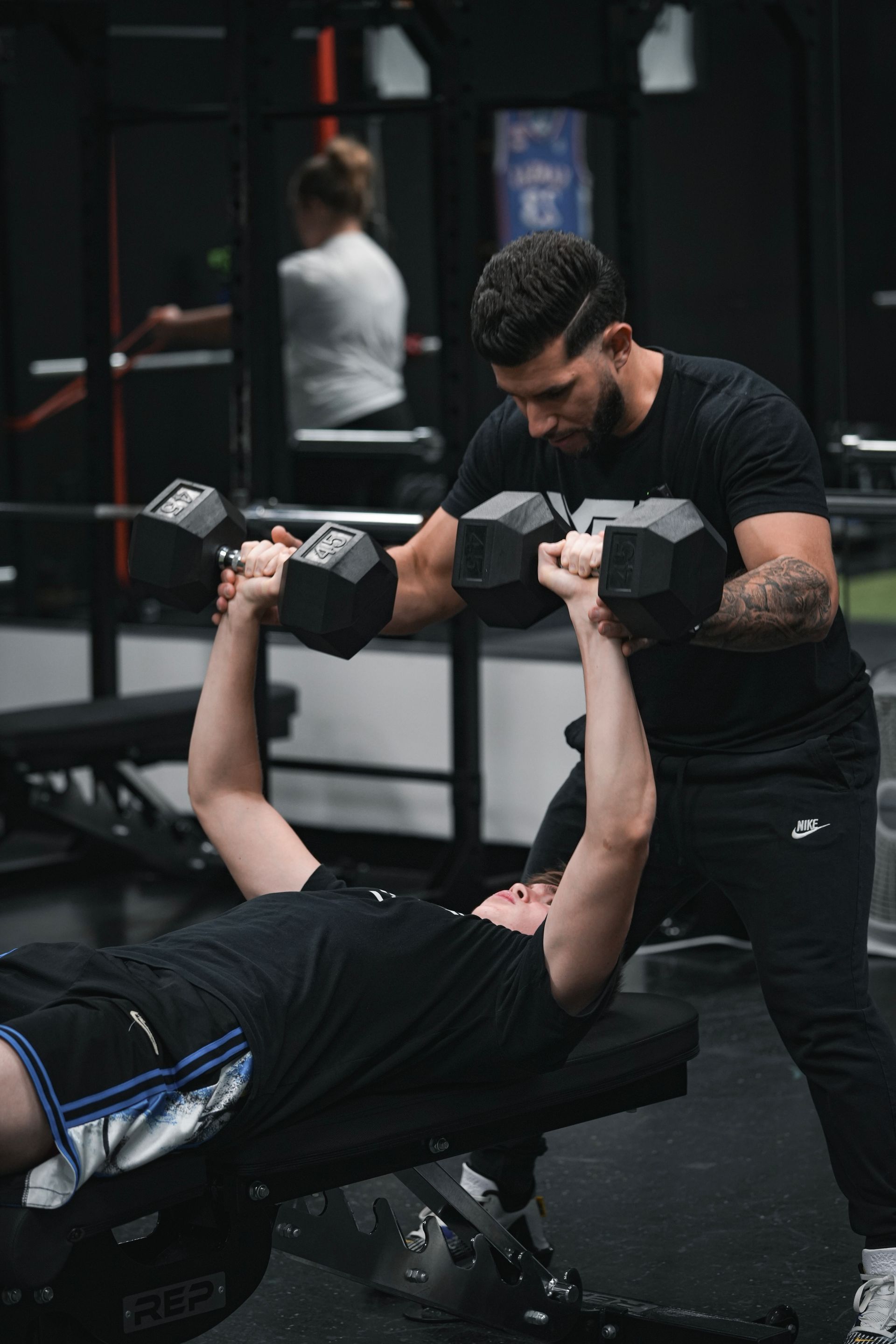A man is helping another man lift dumbbells in a gym.