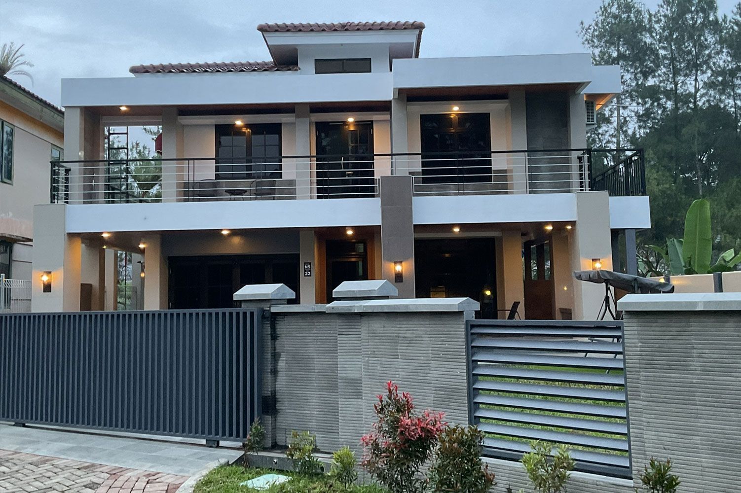 Two-story house with balcony, gray fence, and plants in front.