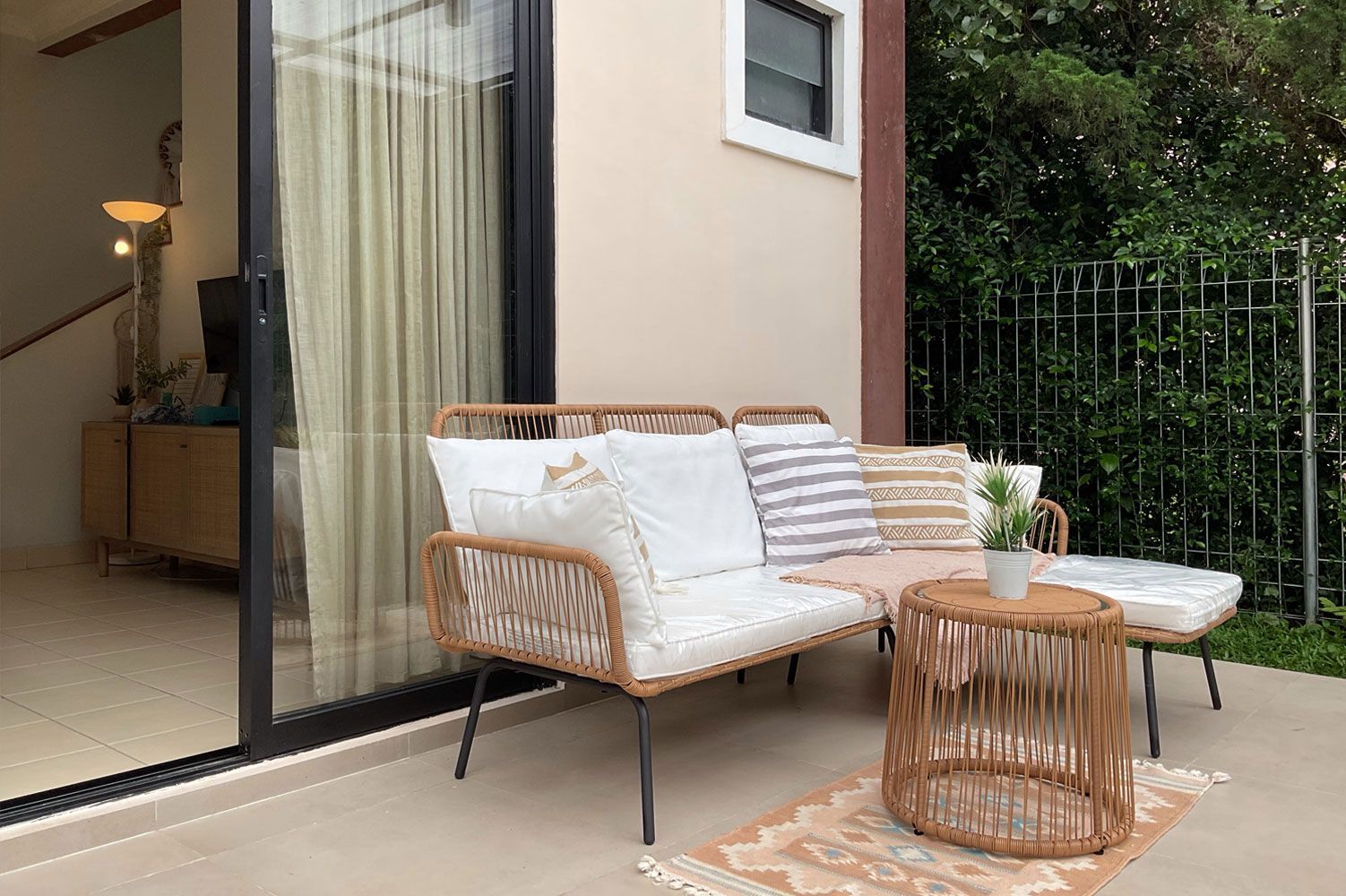 Patio with seating: rattan sofa, cushions, small table, rug, greenery.