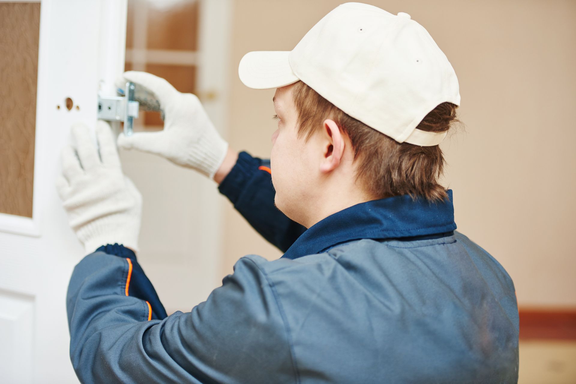 Male carpenter wearing blue uniform installs a lock on a white door. Male carpenter wearing blue uniform installs a lock on a white door.