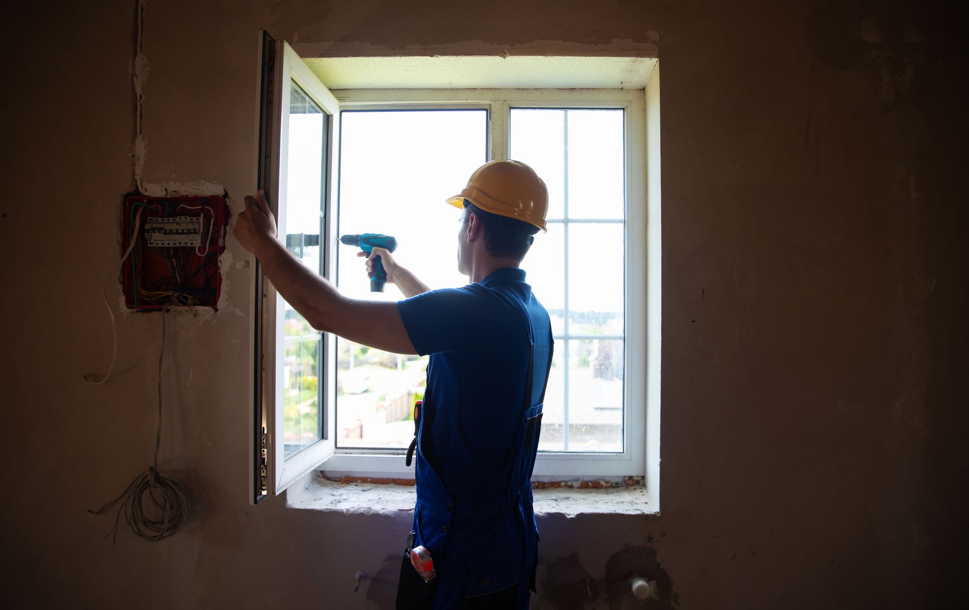 A male contractor in a yellow hard hat is working on window replacement, with a drill on hands.