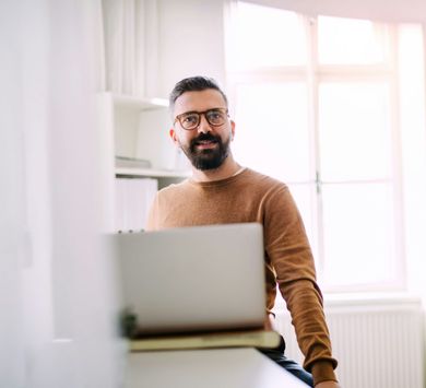 Man with beard and glasses smiles, leaning on a desk with a laptop, in front of a window.