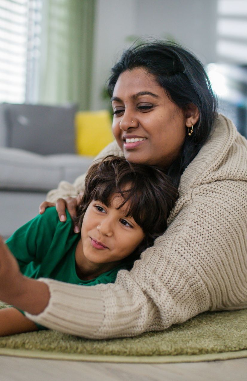 Woman and boy lying on rug, taking selfie. Woman smiles. Boy looks at phone. Soft light indoors.