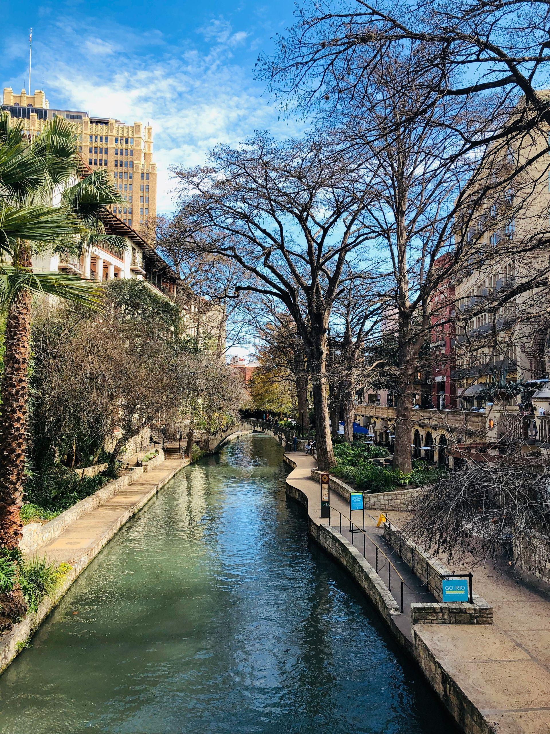San Antonio River Walk: a waterway lined with stone pathways, restaurants, and trees, under a blue sky.
