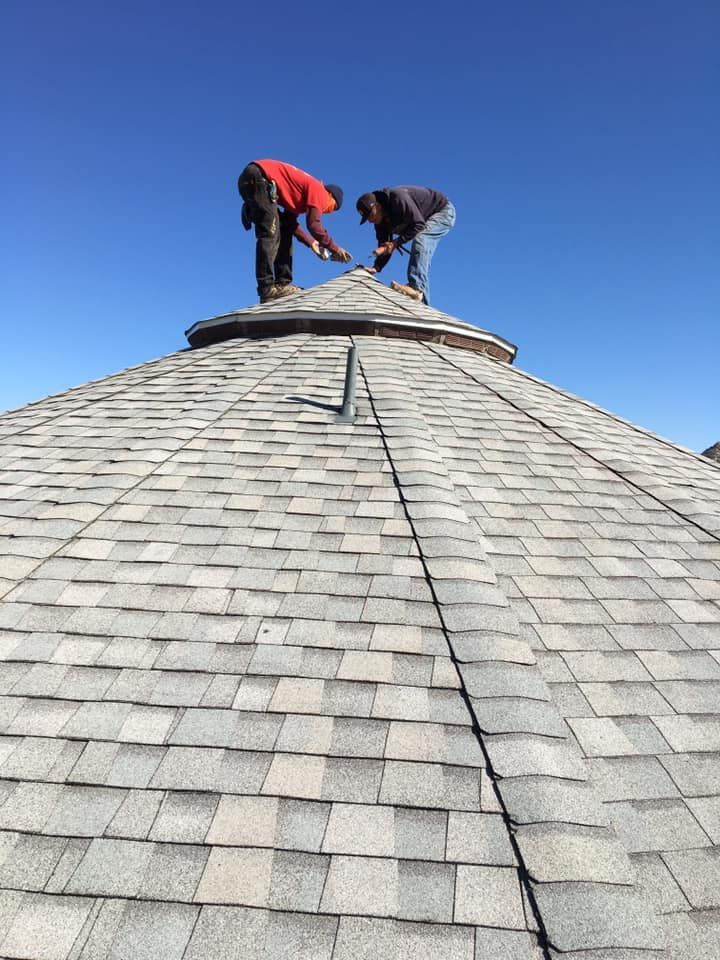 Two roofers working on a shingled, conical roof under a clear blue sky.