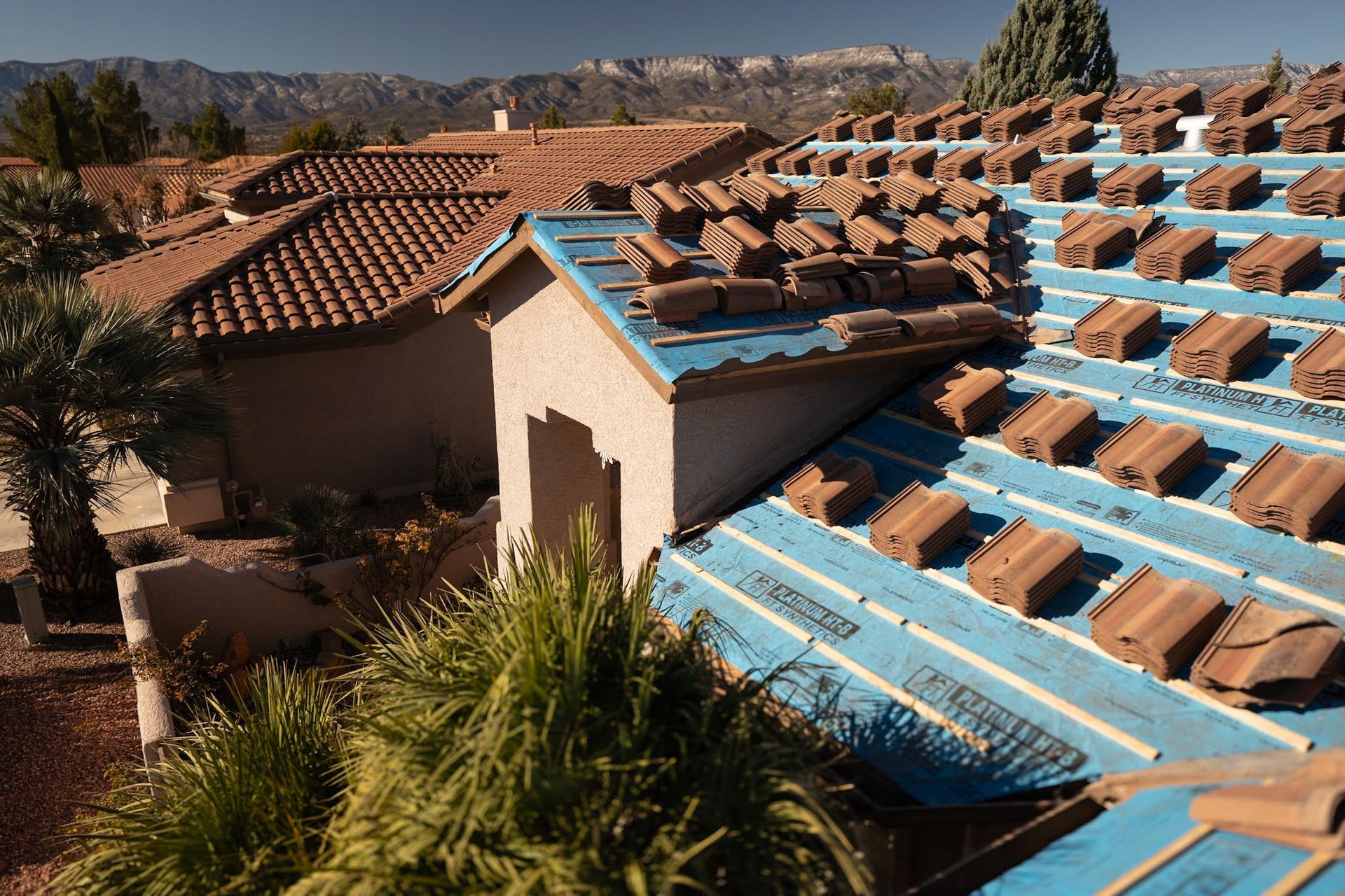 Roof partially covered with terracotta tiles; blue underlayment exposed, houses in background.