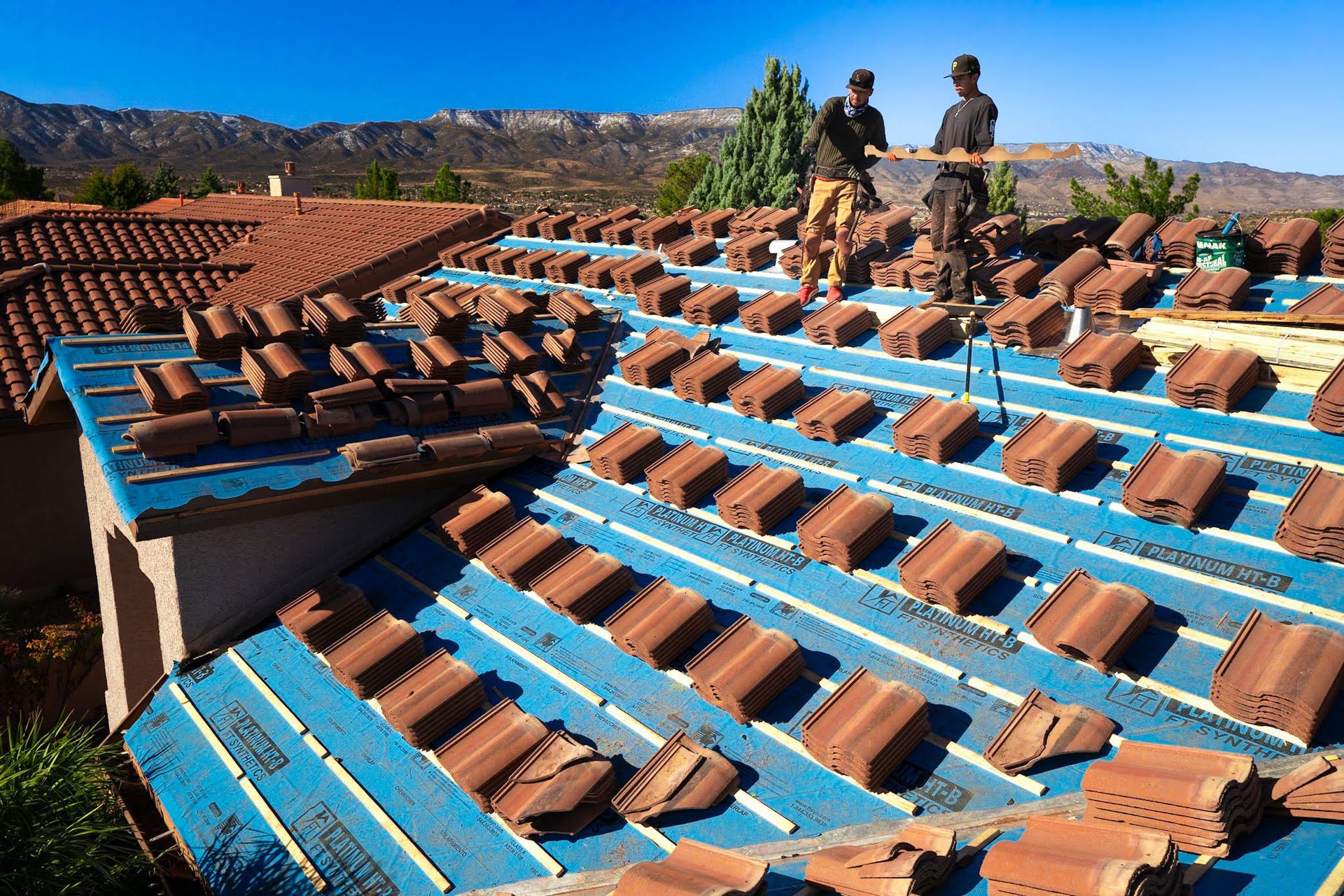Two roofers installing terra cotta tiles on a blue underlayment. Mountains in the background.