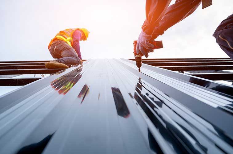 Construction workers installing a metal roof, one drilling, sunny sky.