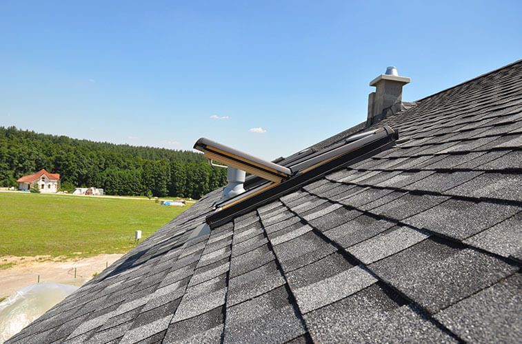 Roof with open skylight, chimney, and grey shingles against a blue sky, with a green field and trees in the background.