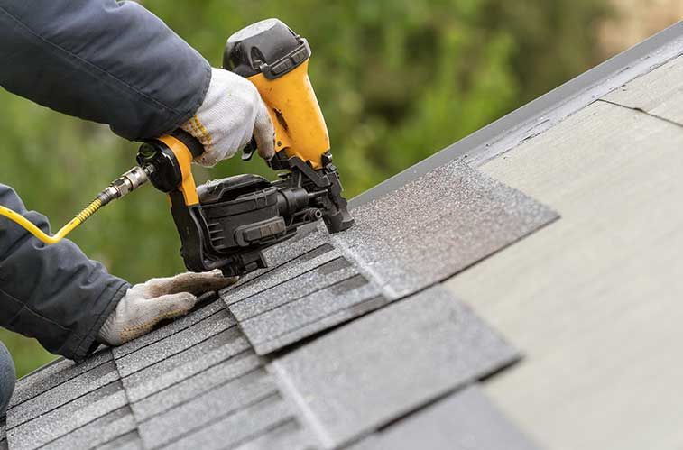 Roofer using a nail gun to install asphalt shingles on a roof, wearing gloves and a jacket.