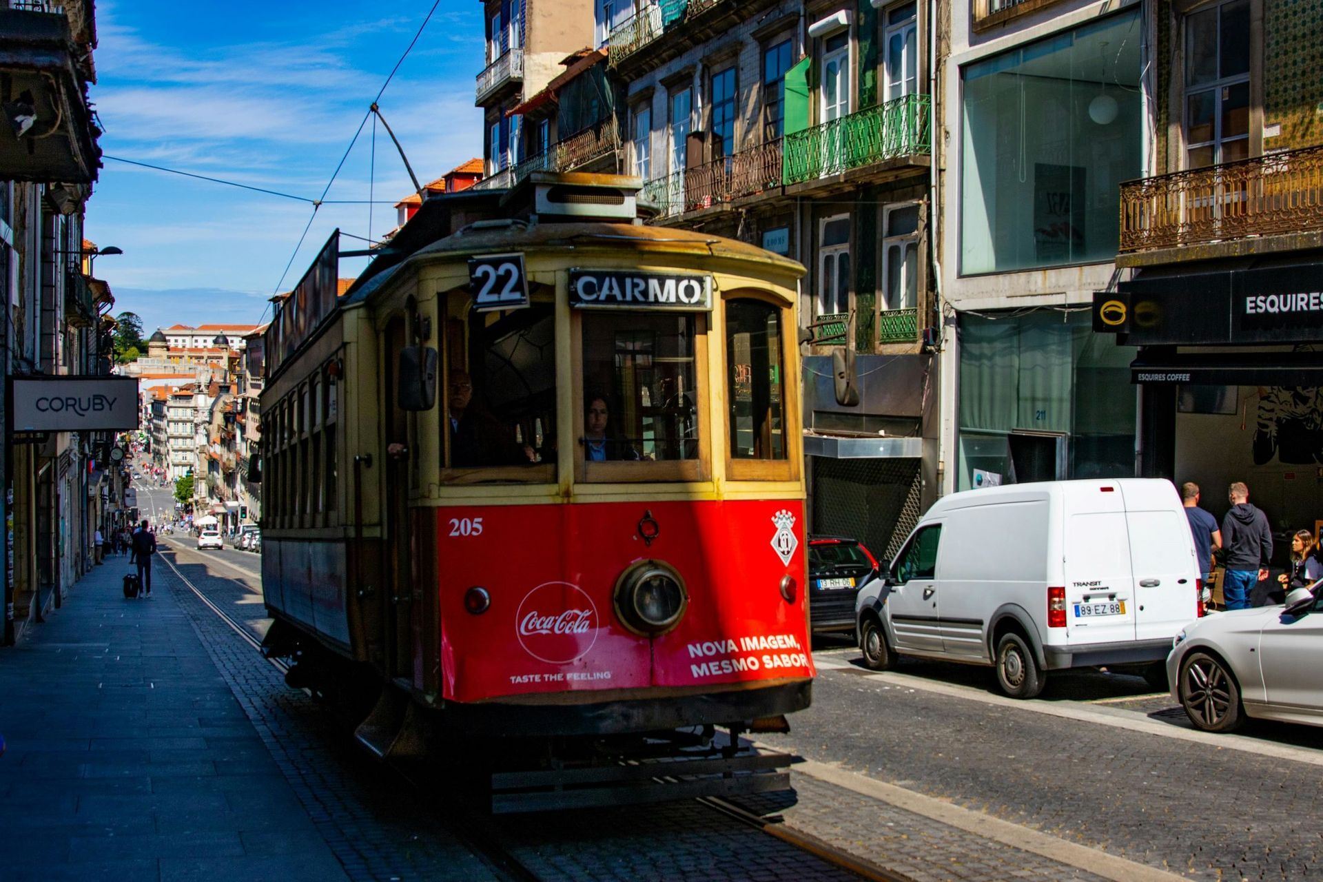 Red and yellow tram on tracks in a narrow street, lined with buildings and parked cars.