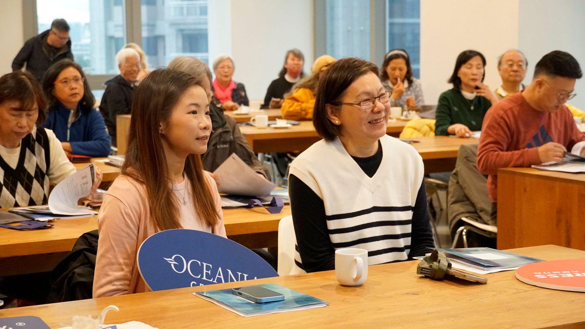 人們圍坐在房間裡的桌旁，參加會議或講座。有些人面帶微笑，認真聆聽。