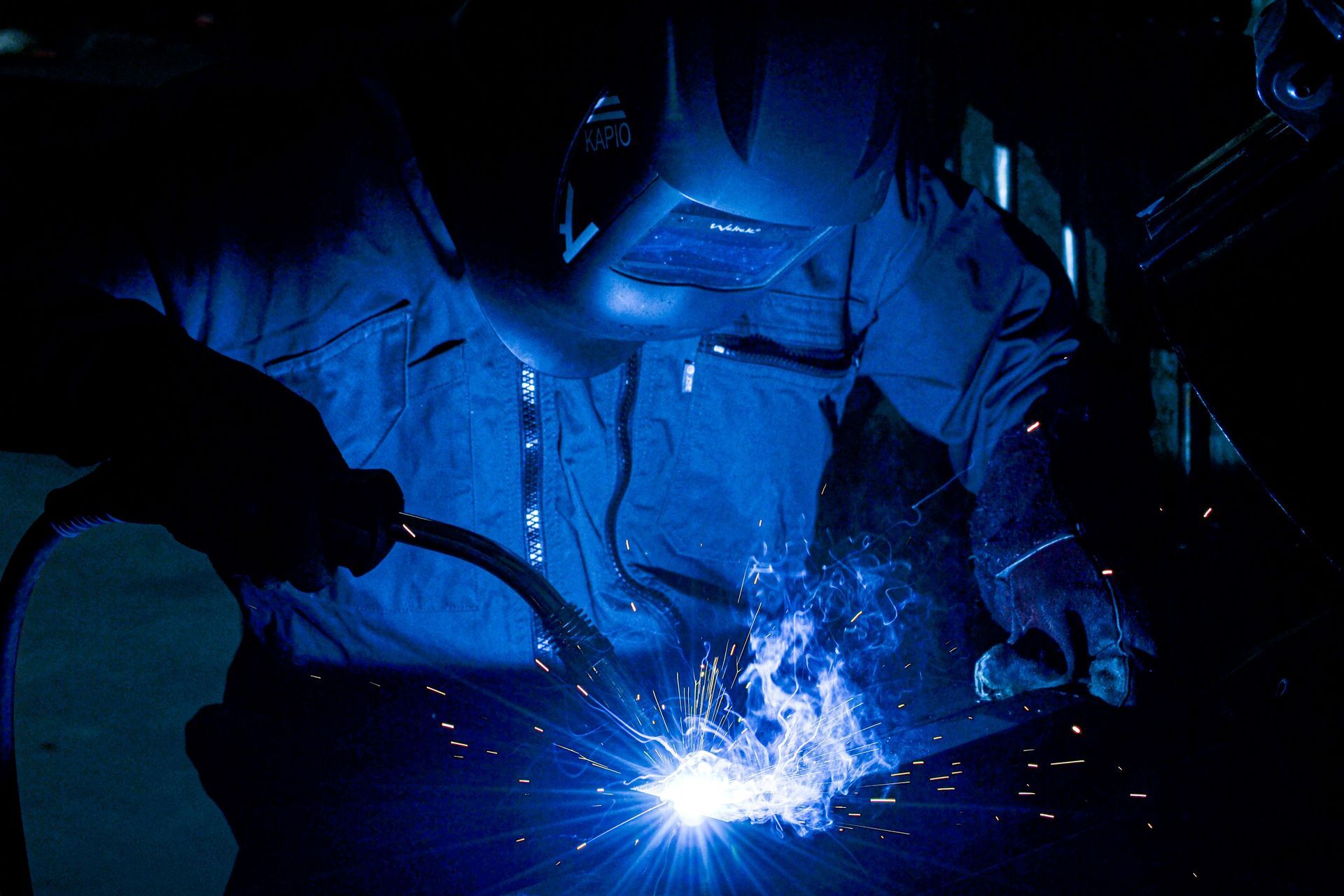Welder in blue light sparks at a metalwork bench, wearing protective gear and helmet.