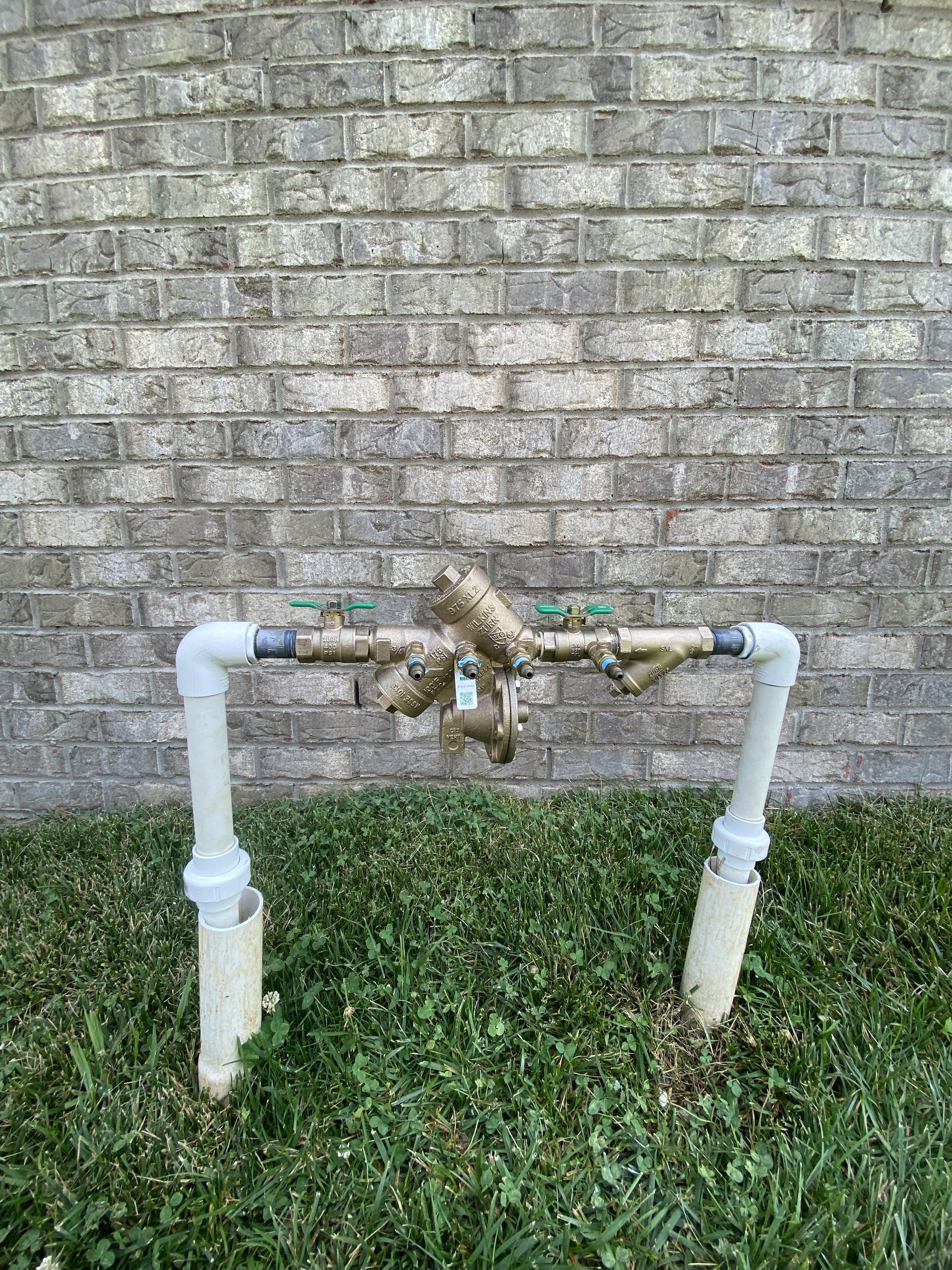 Water pipes with valves in front of a brick wall, surrounded by grass.