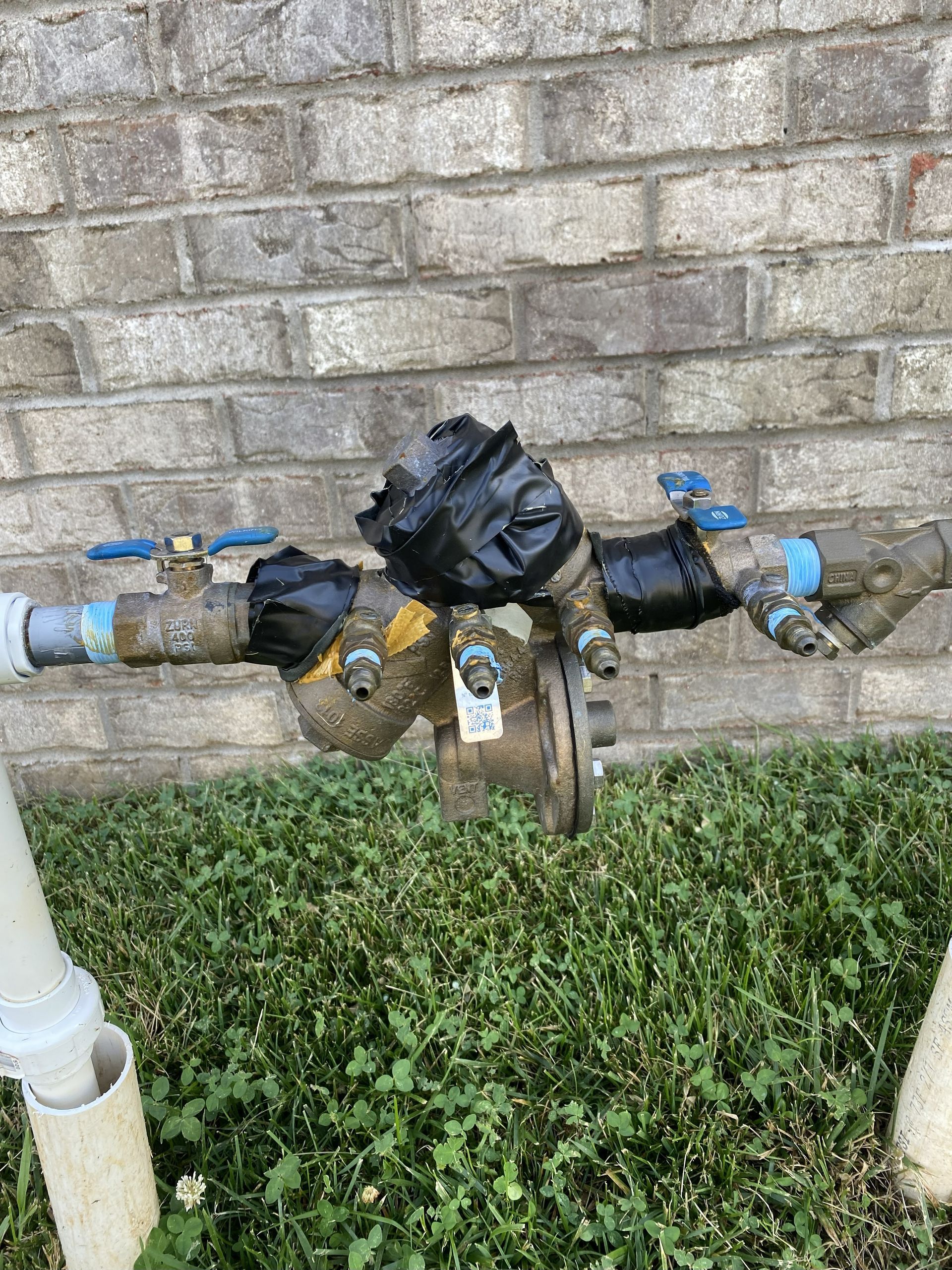 Water pipes with valves, wrapped in black tape, against a brick wall. Green grass in foreground.