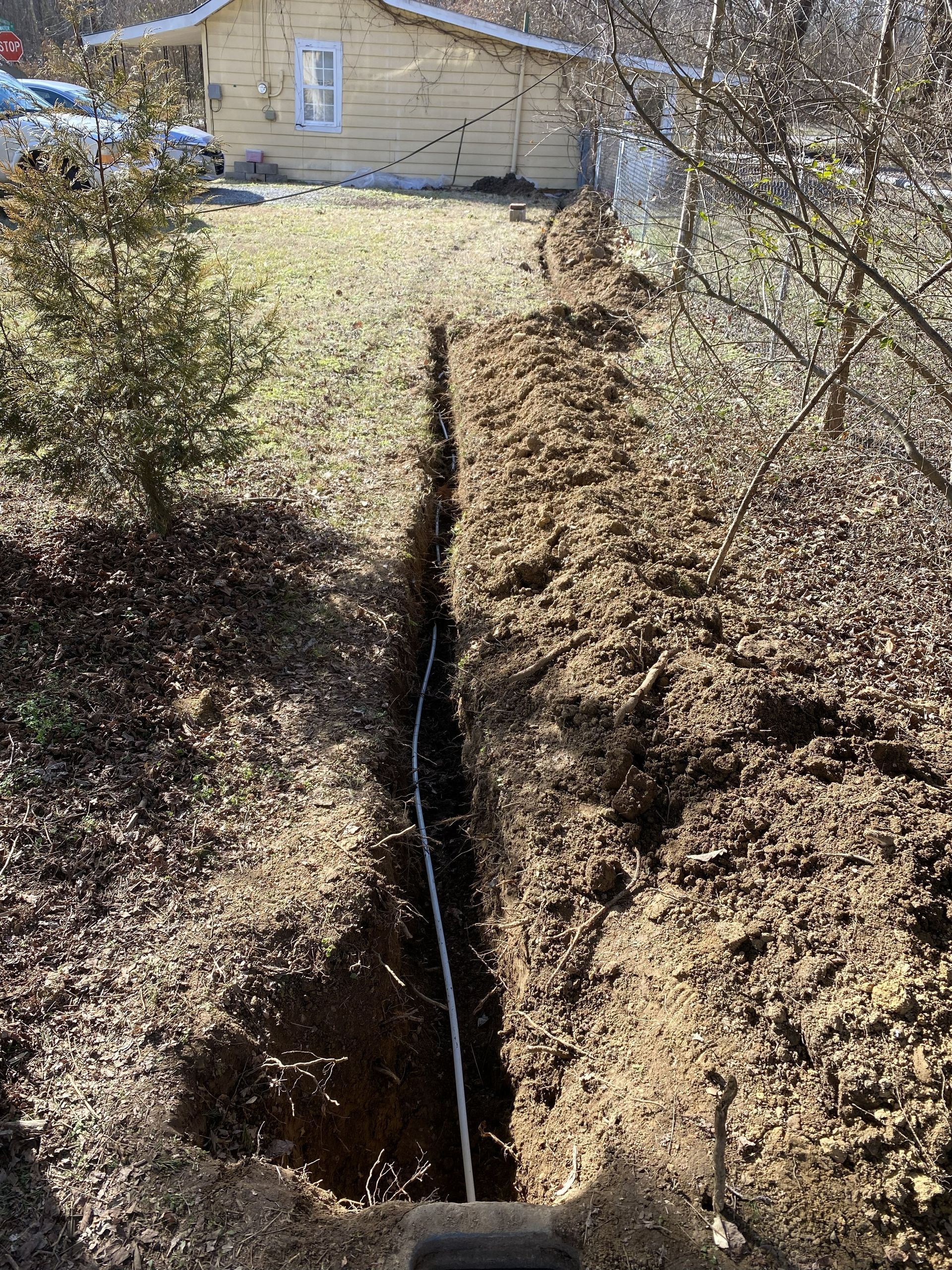 A narrow trench dug in the dirt with a white pipe inside, leading to a yellow house in the background.