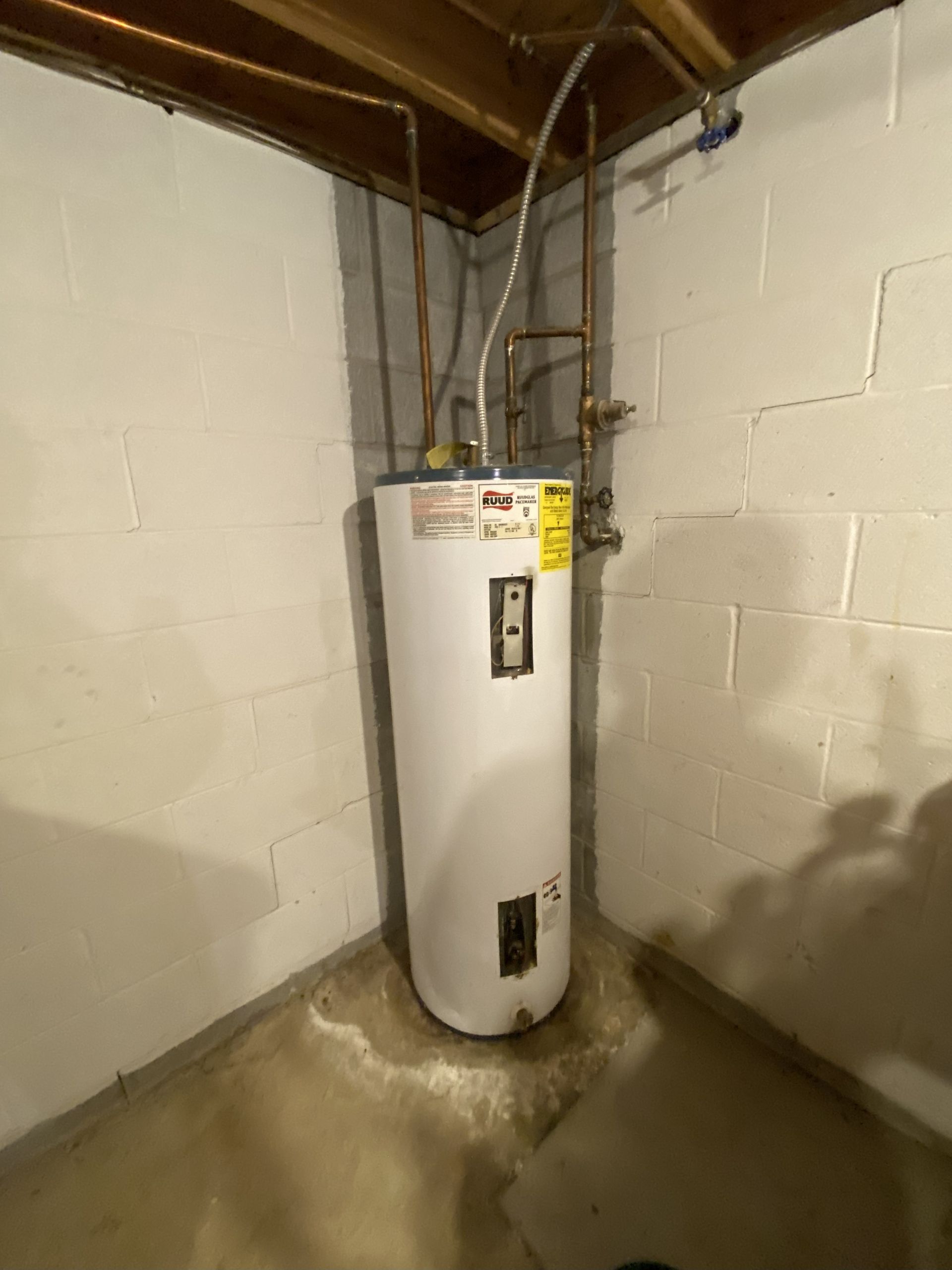 Water heater in a basement corner against white block walls. Copper pipes and water stain on the concrete floor.