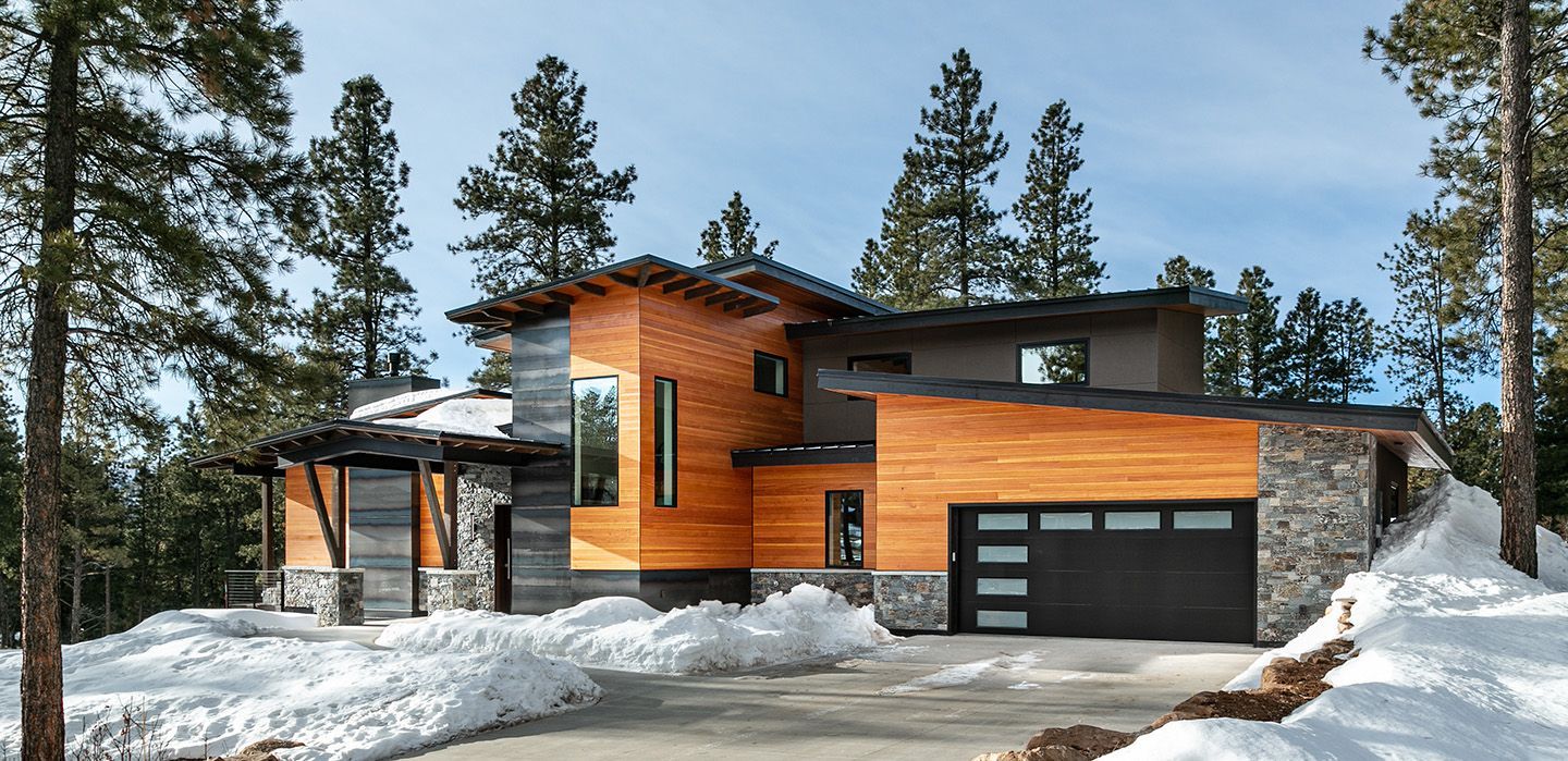 A house with a garage is surrounded by snow and trees.