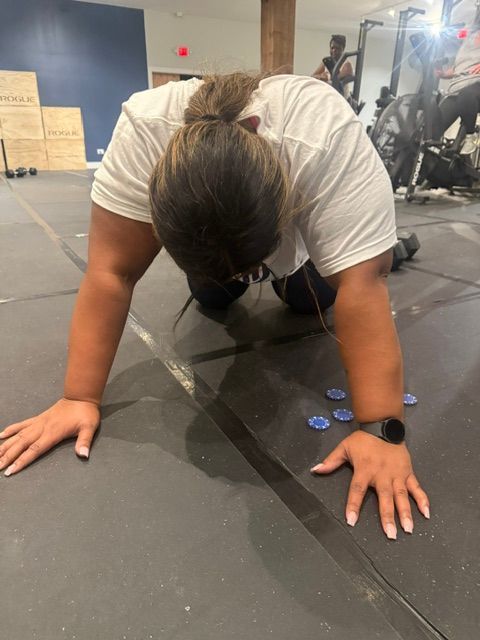 Woman bent over on floor, hands out, possibly resting after exercise; blue and grey gym setting.