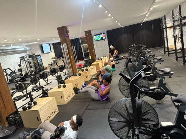 People exercising in a gym, using weights and exercise bikes. Well-lit interior with black and wooden accents.