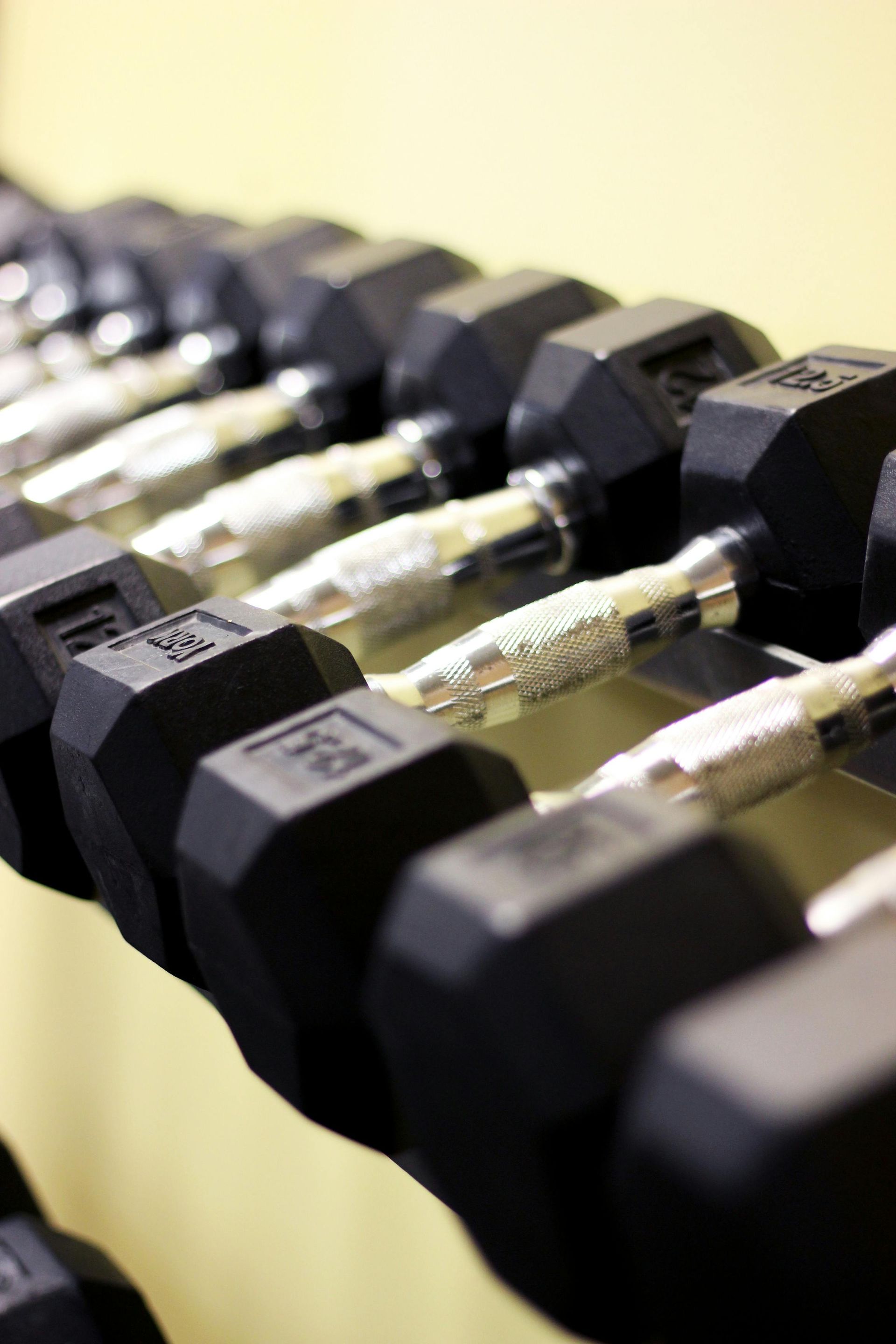 Row of black dumbbells on a rack, metal handles, set against a light yellow wall.