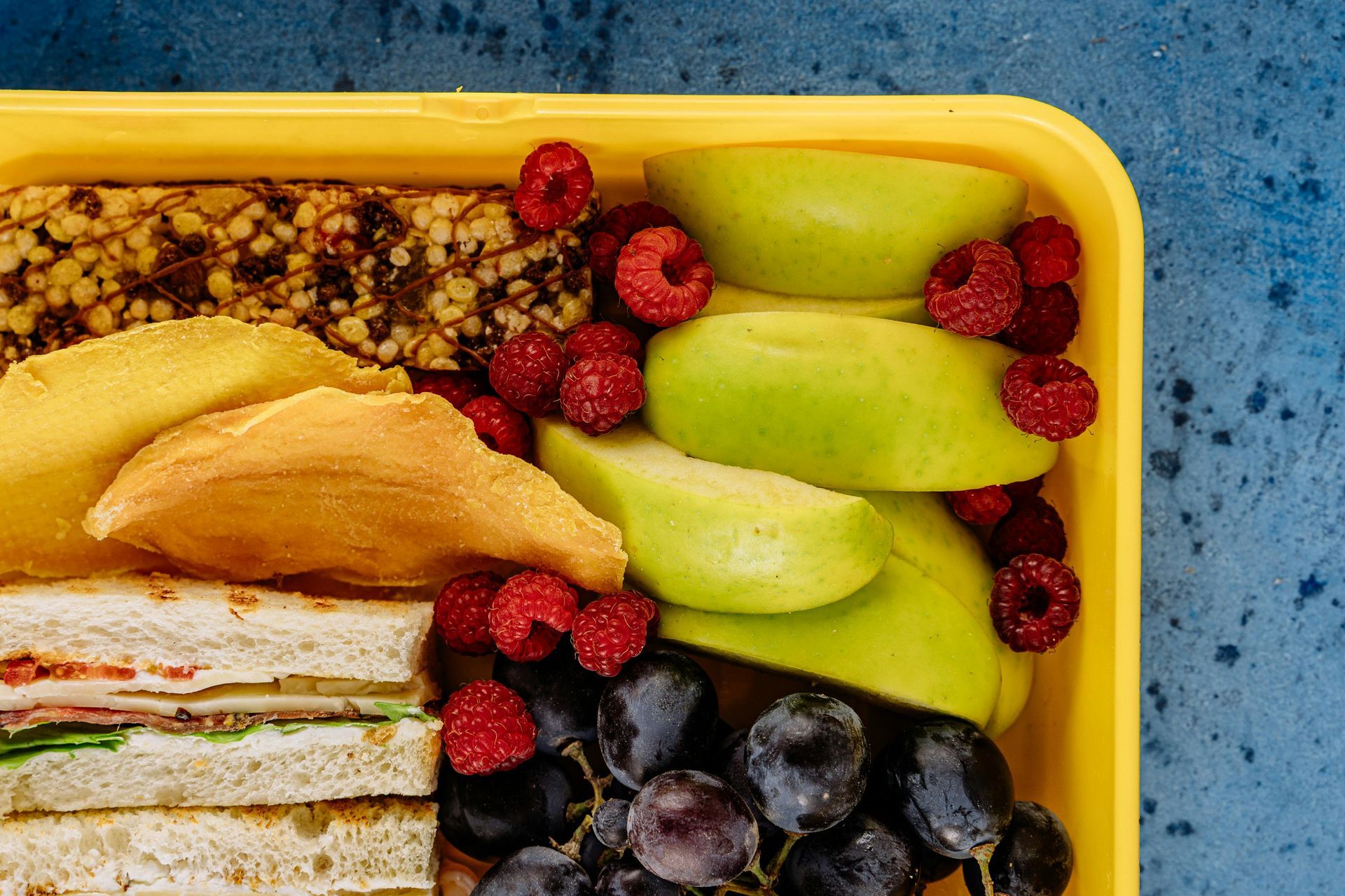 Yellow lunchbox with sandwich, bar, apple slices, raspberries, and grapes.