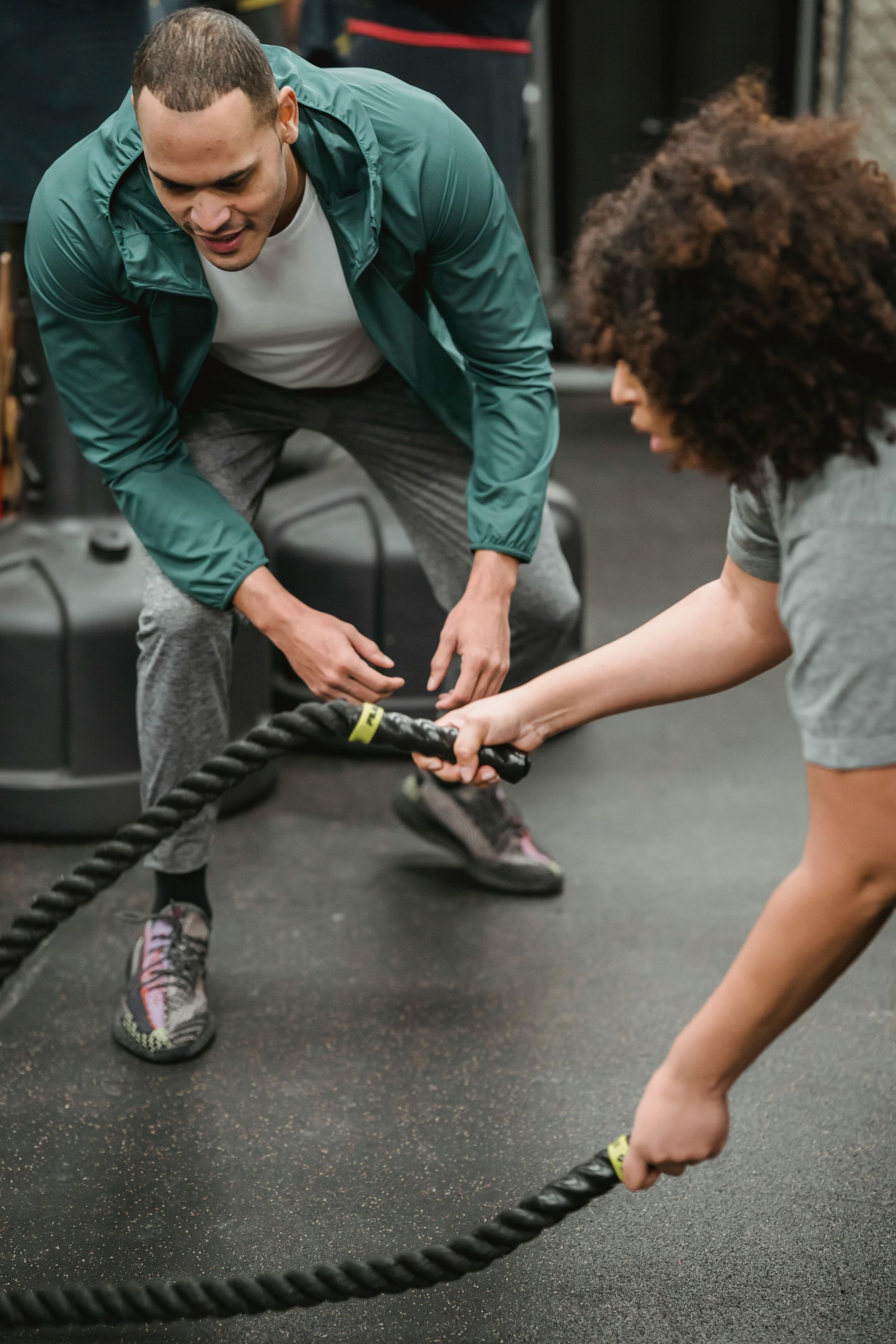 Man guiding another using battle ropes in a gym. Green jacket, smiling, dark hair, and gray floor.