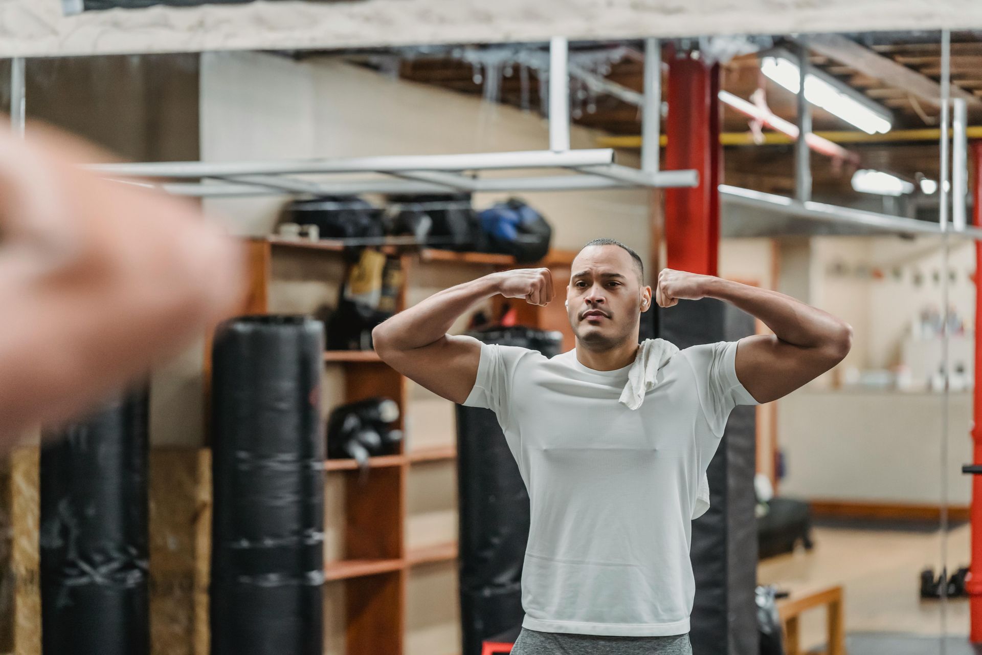 Man flexes biceps in gym mirror, white shirt, towel, neutral expression.