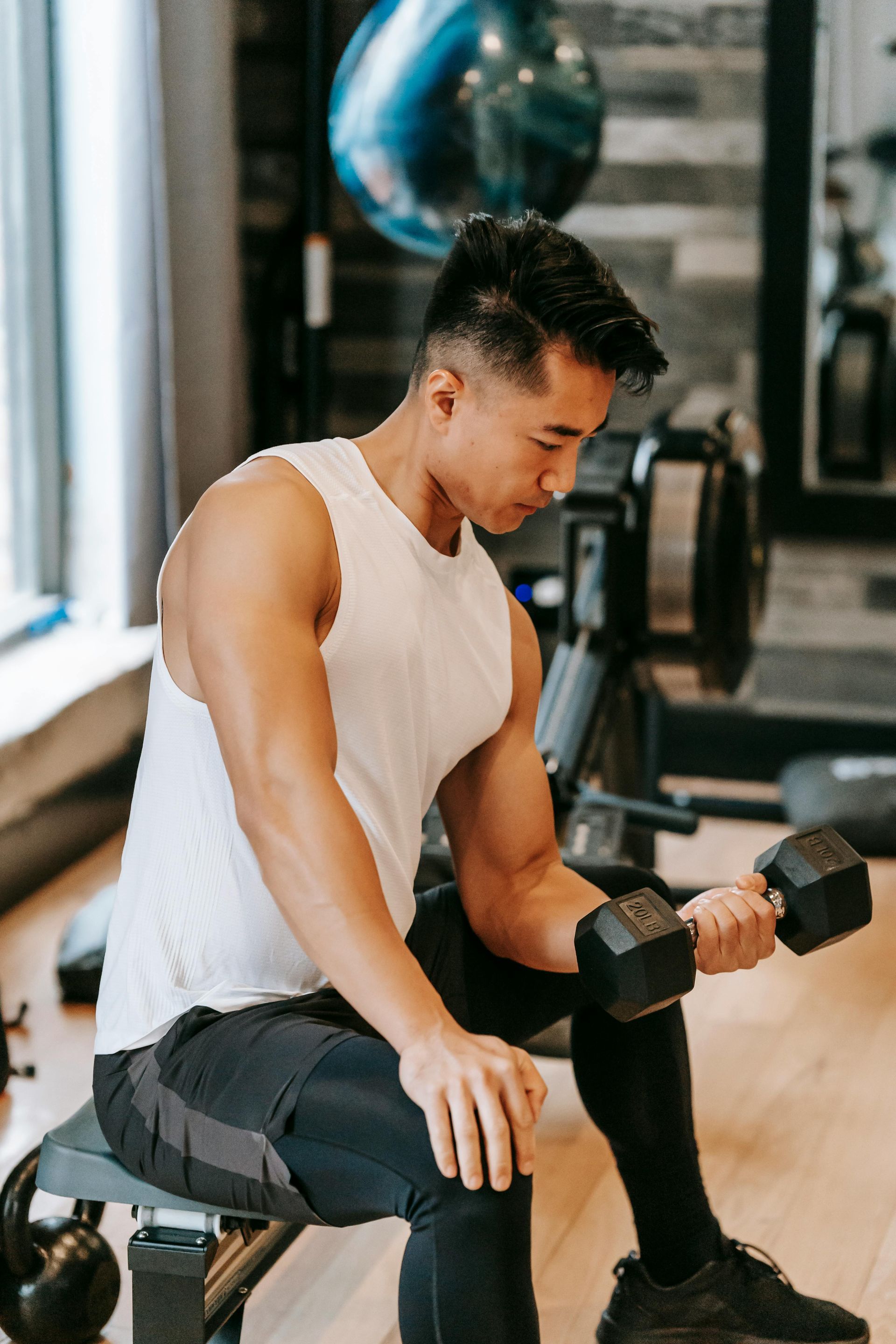 Man in a gym curls a dumbbell while seated on a bench. He wears a white tank top and black workout pants.