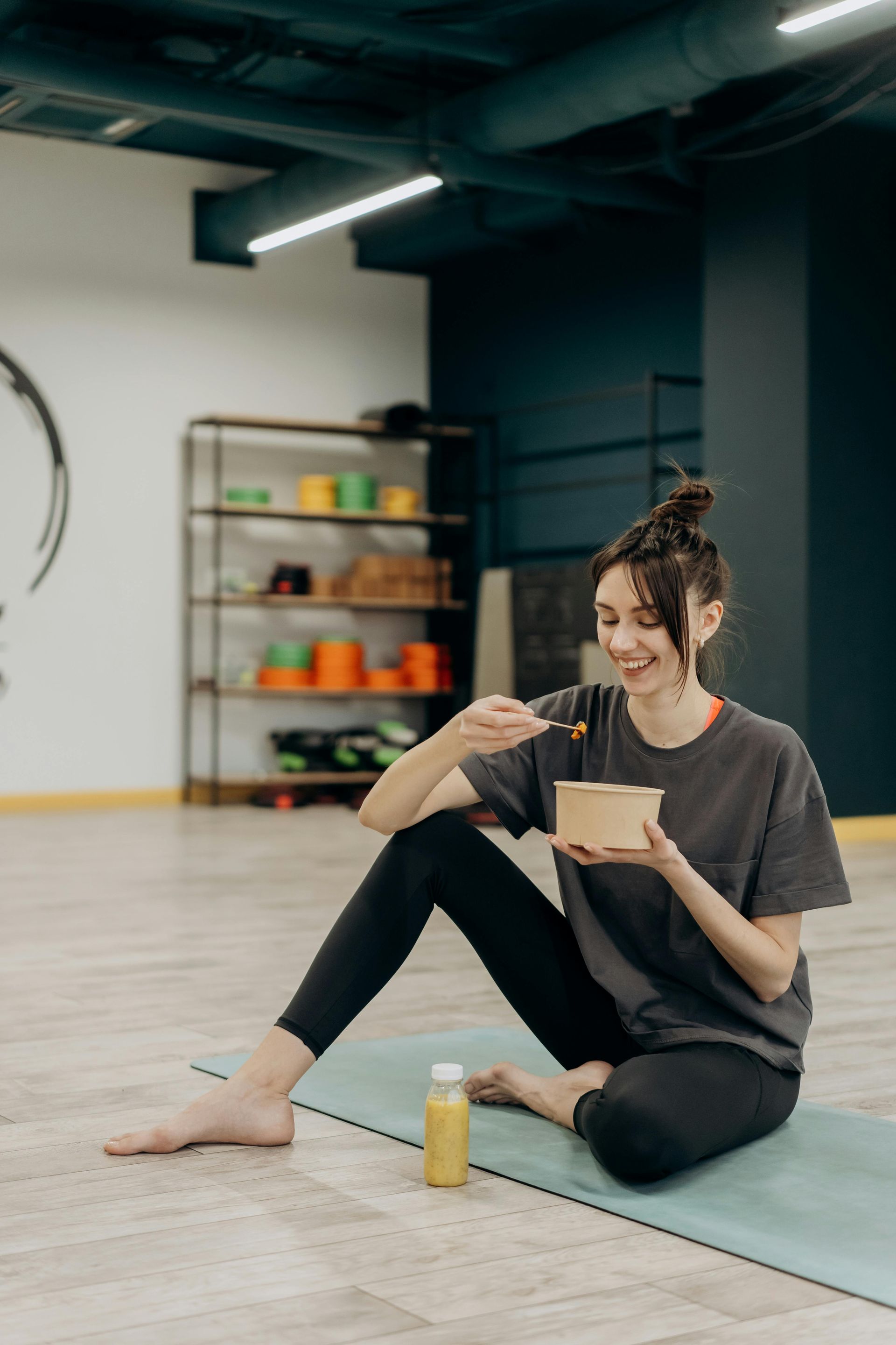 Woman in workout clothes eating from a bowl on a yoga mat, bottle beside her, in a studio.