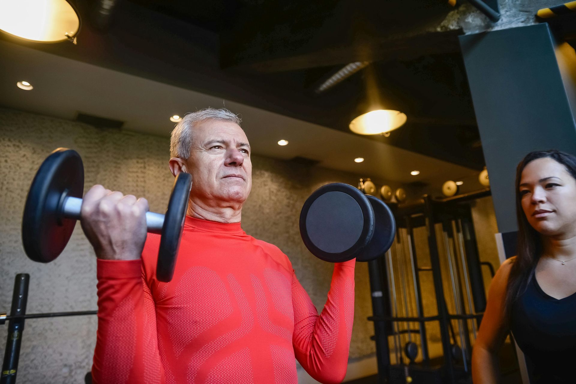 Man in red shirt lifting dumbbells at gym, woman watching.