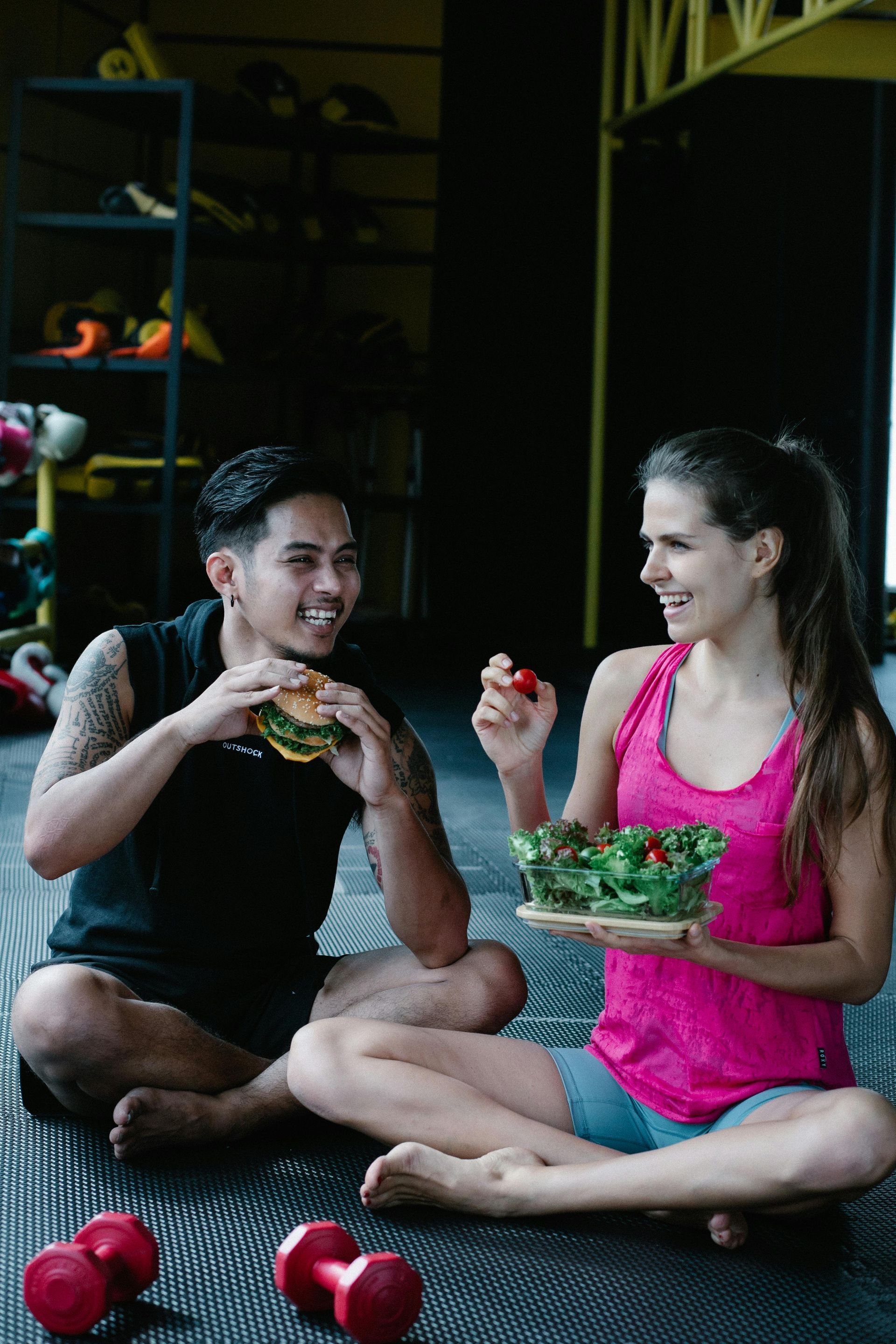 Man eating a burger, woman eating a salad, sitting on gym floor, smiling.