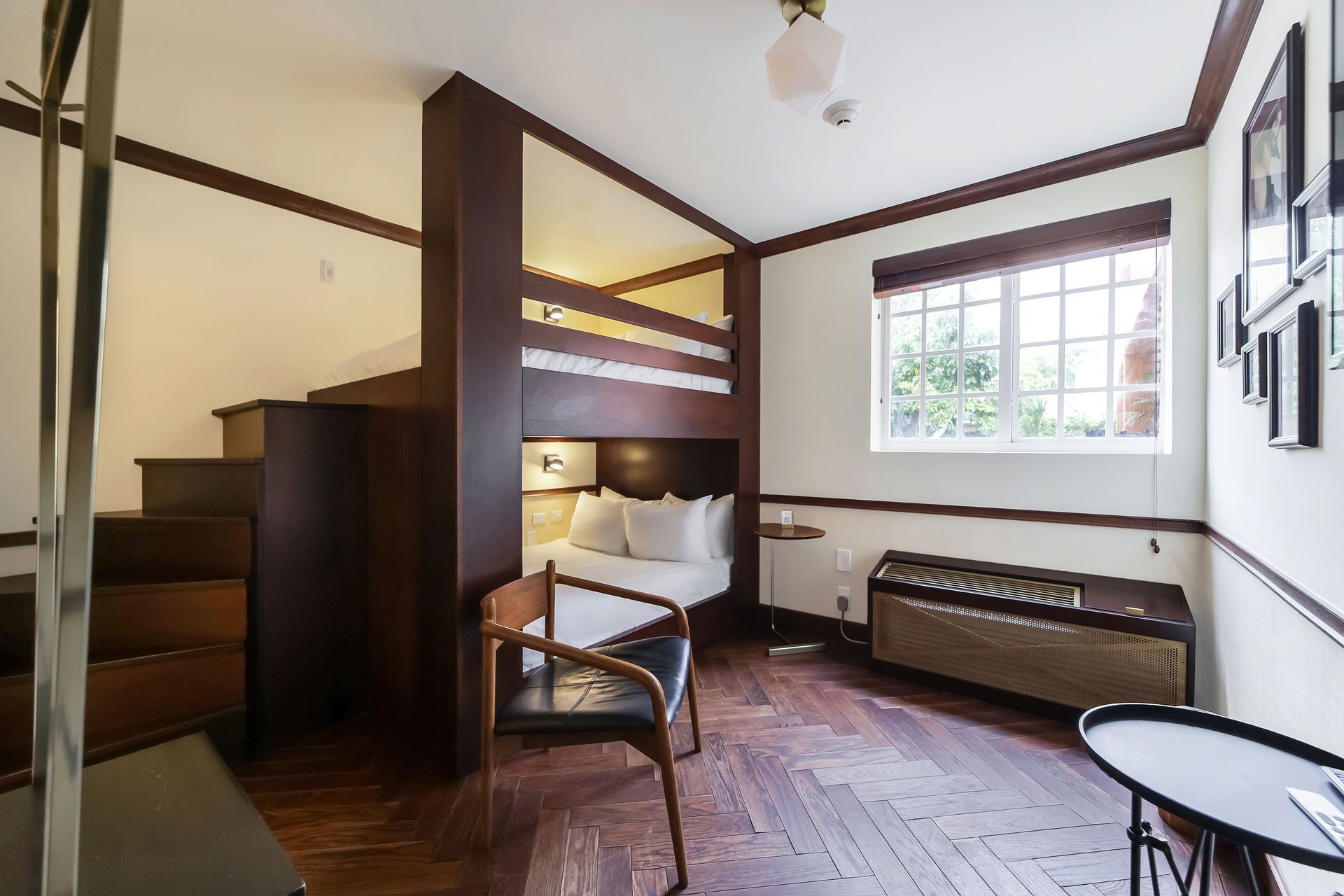 A small hotel room with a bunk bed, a chair, and a window. Wooden floors and dark wood accents.