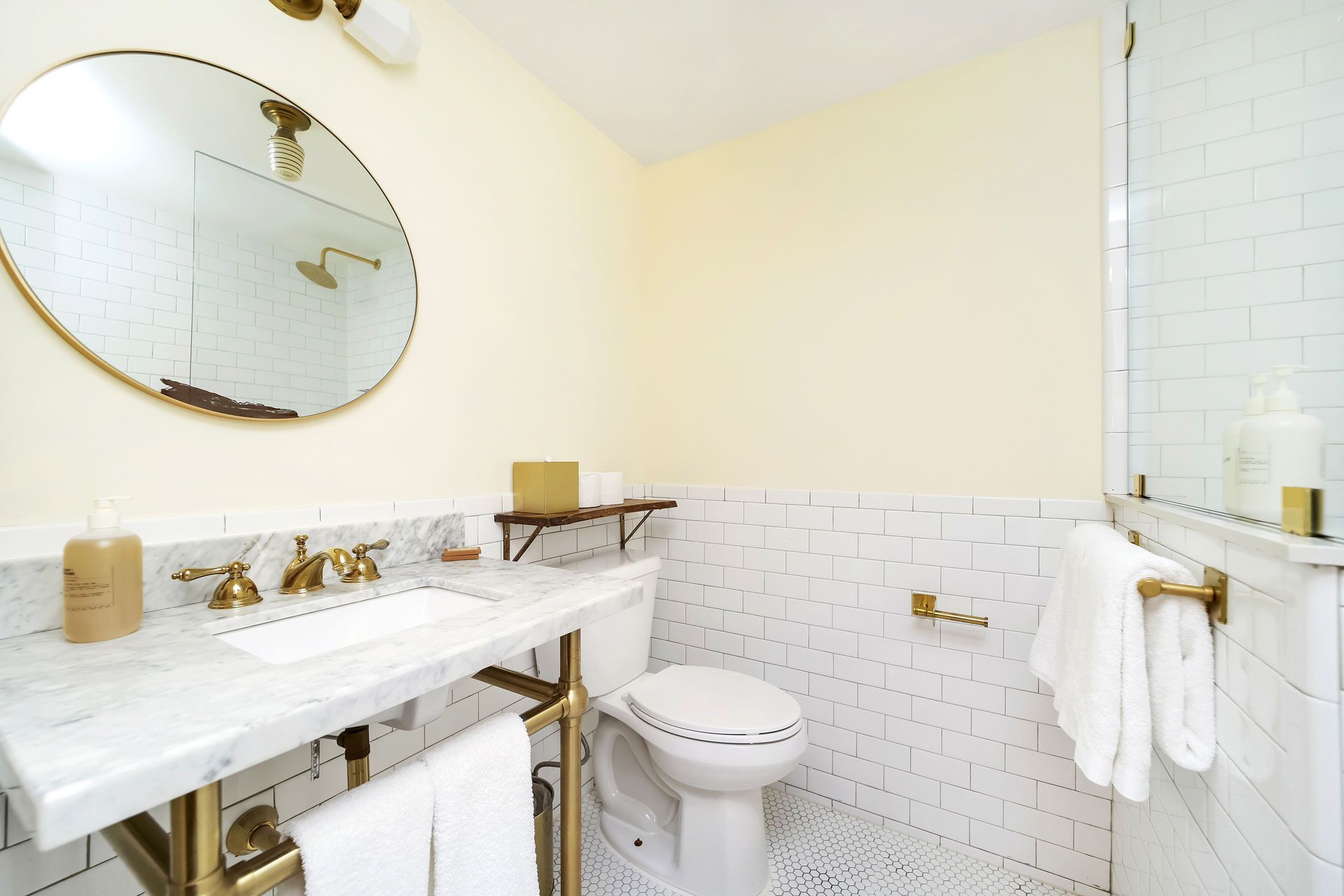 Bathroom with marble vanity, gold fixtures, round mirror, white tile, and yellow walls.