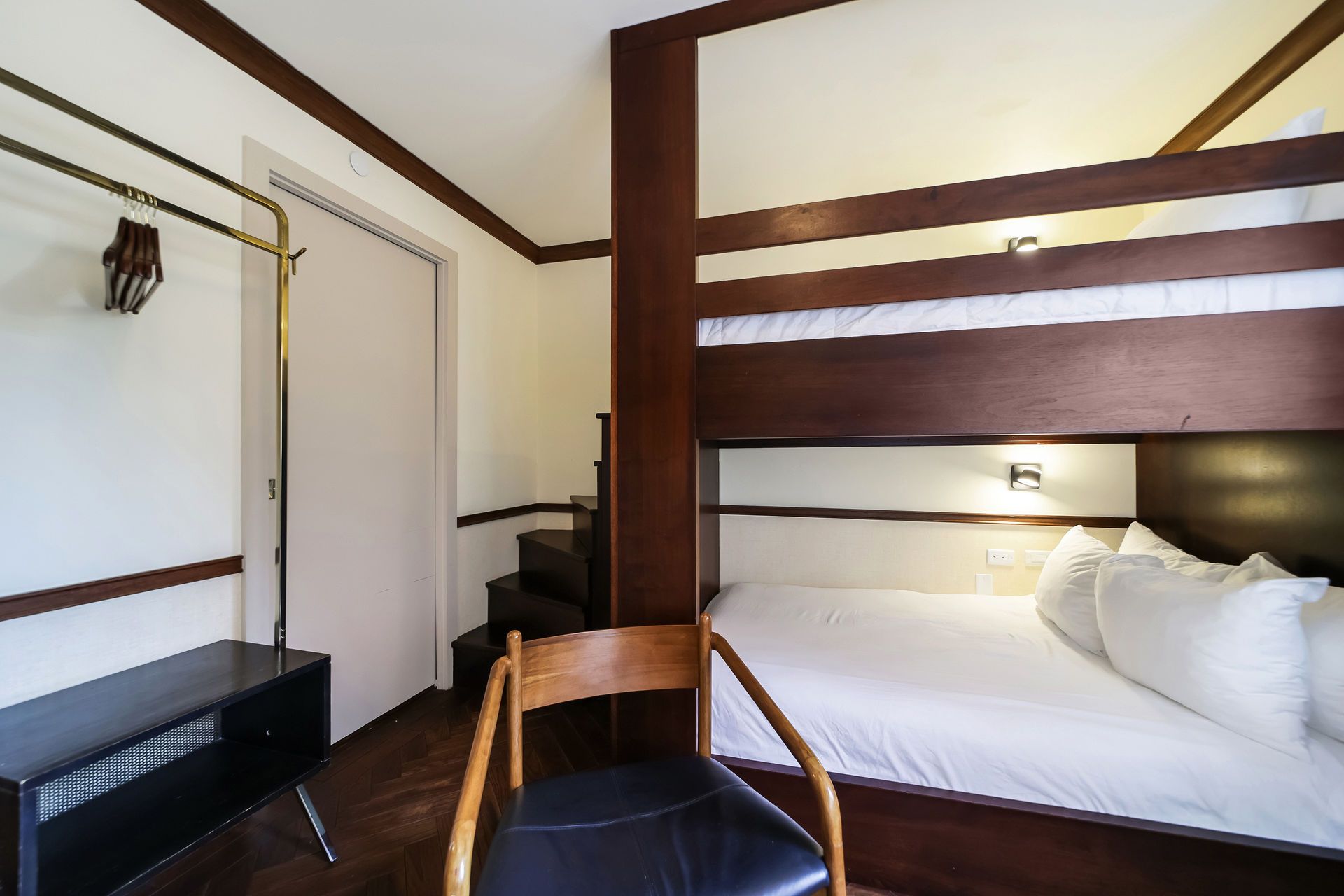 Bunk bed in a room with a black bench, chair, and clothes rack. White sheets, dark wood.