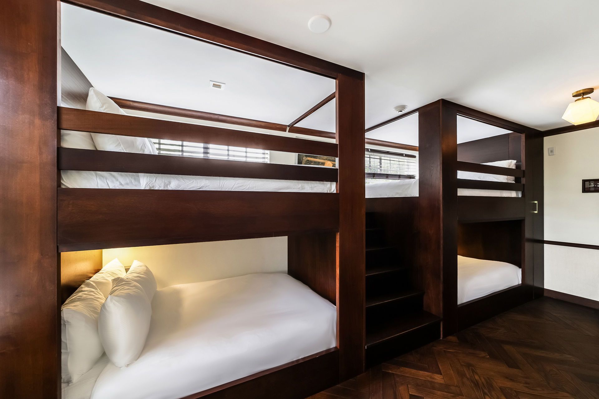 Dark wood bunk beds in a room; each has a bottom and top bed. Wooden floor and white bedding.
