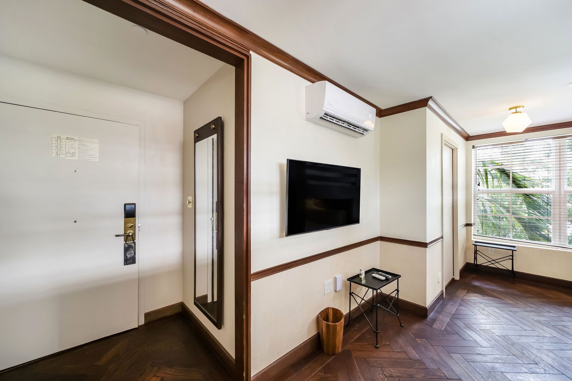 Interior room with door, mirror, TV, window, and wood floor. Walls are cream with dark wood trim.