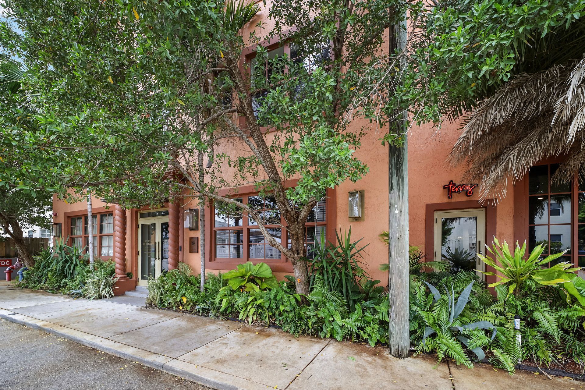 Exterior of a coral-colored building with trees and lush greenery. Includes a thatched roof and sidewalk.