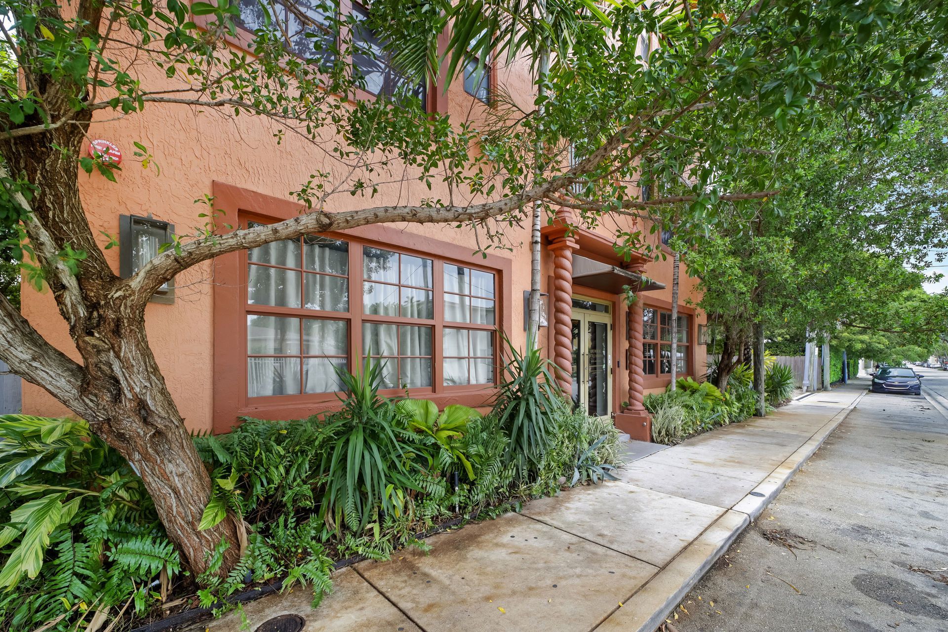 Building with orange stucco facade, brown trim, large windows, and lush greenery along the sidewalk.