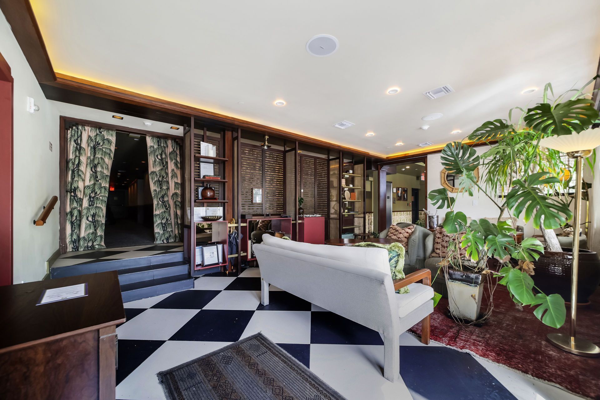 Black and white checkered floor in a room. A white couch sits in the center. Large plants and open doorway visible.