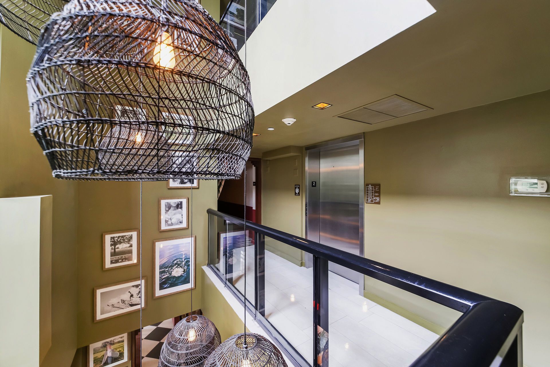 Hallway with elevator, black wicker light fixtures, glass railing, and framed artwork on the wall.
