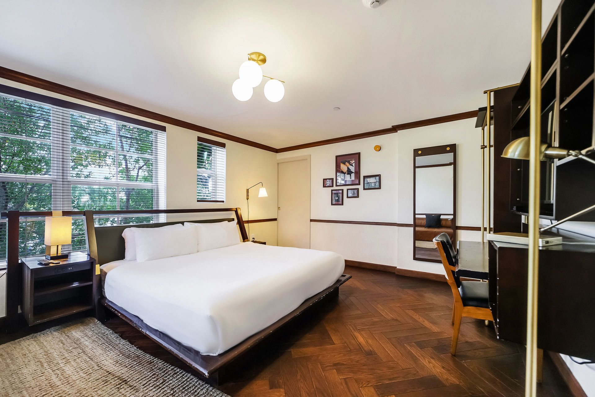 Hotel bedroom with a bed, desk, and large window overlooking trees. Neutral colors, modern design.
