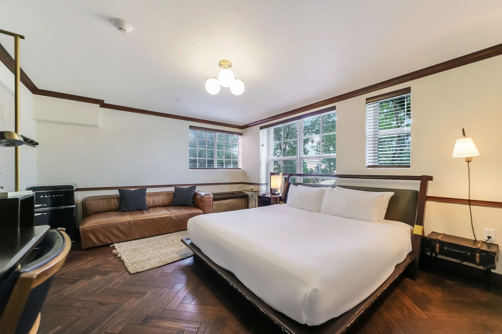 Bedroom with a king bed, brown sofa, and dark wood floors, lit by natural light from windows.