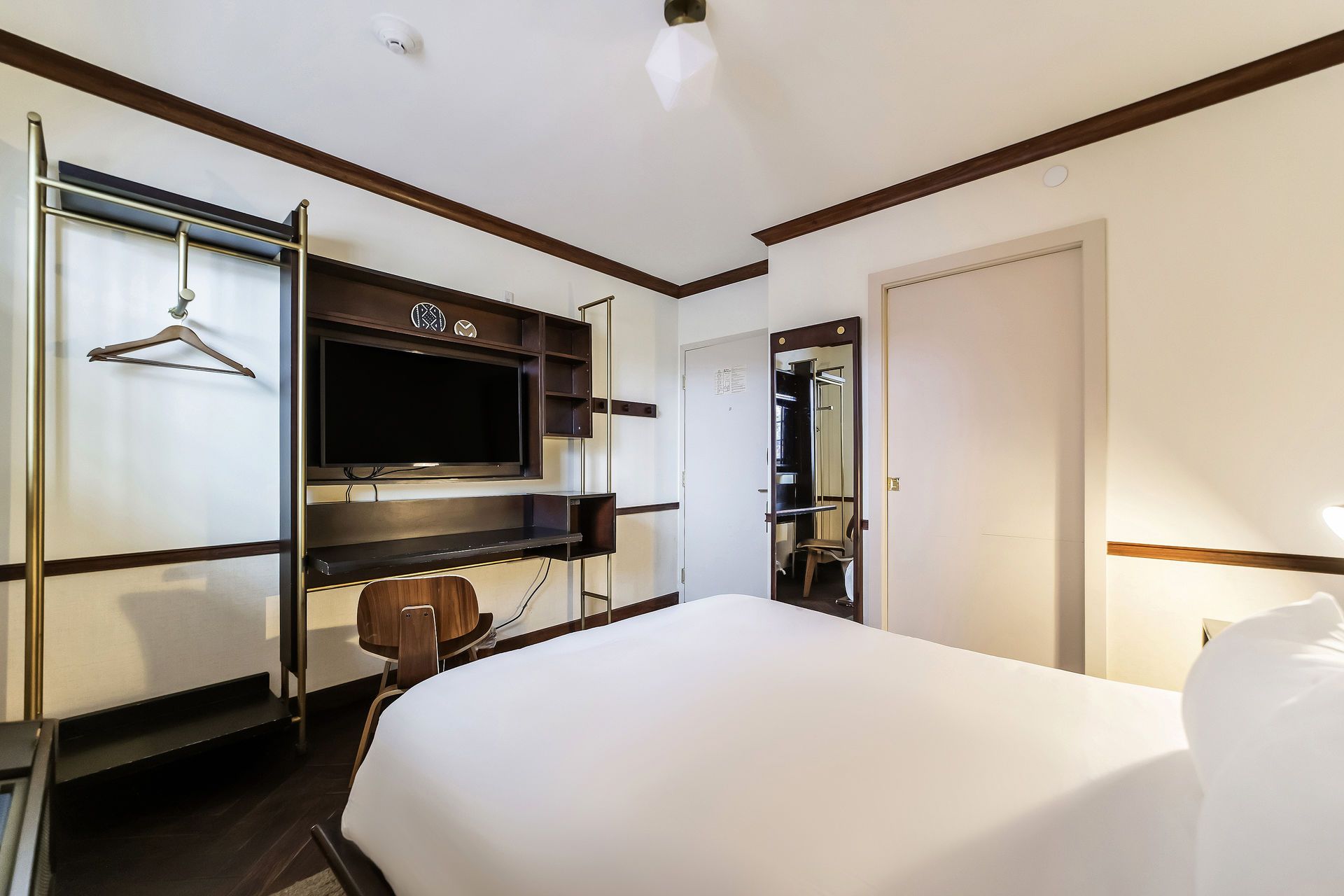 Hotel room with a bed, TV, desk, closet, and a partially visible bathroom. White and brown color scheme.