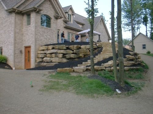 Large house with stone retaining walls, steps, and mulch. Two people on a deck.